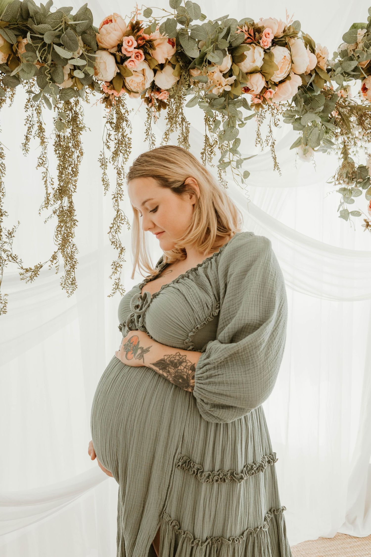A pregnant woman in a long, flowing green dress stands in front of a floral backdrop.