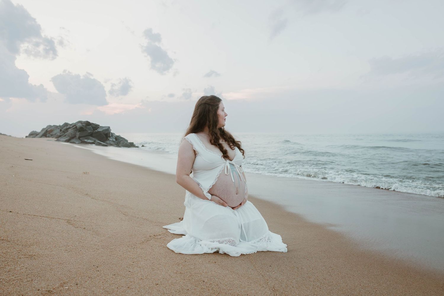 Pregnant woman in white dress sitting on sandy beach at sunset, cradling her baby bump near ocean waves.