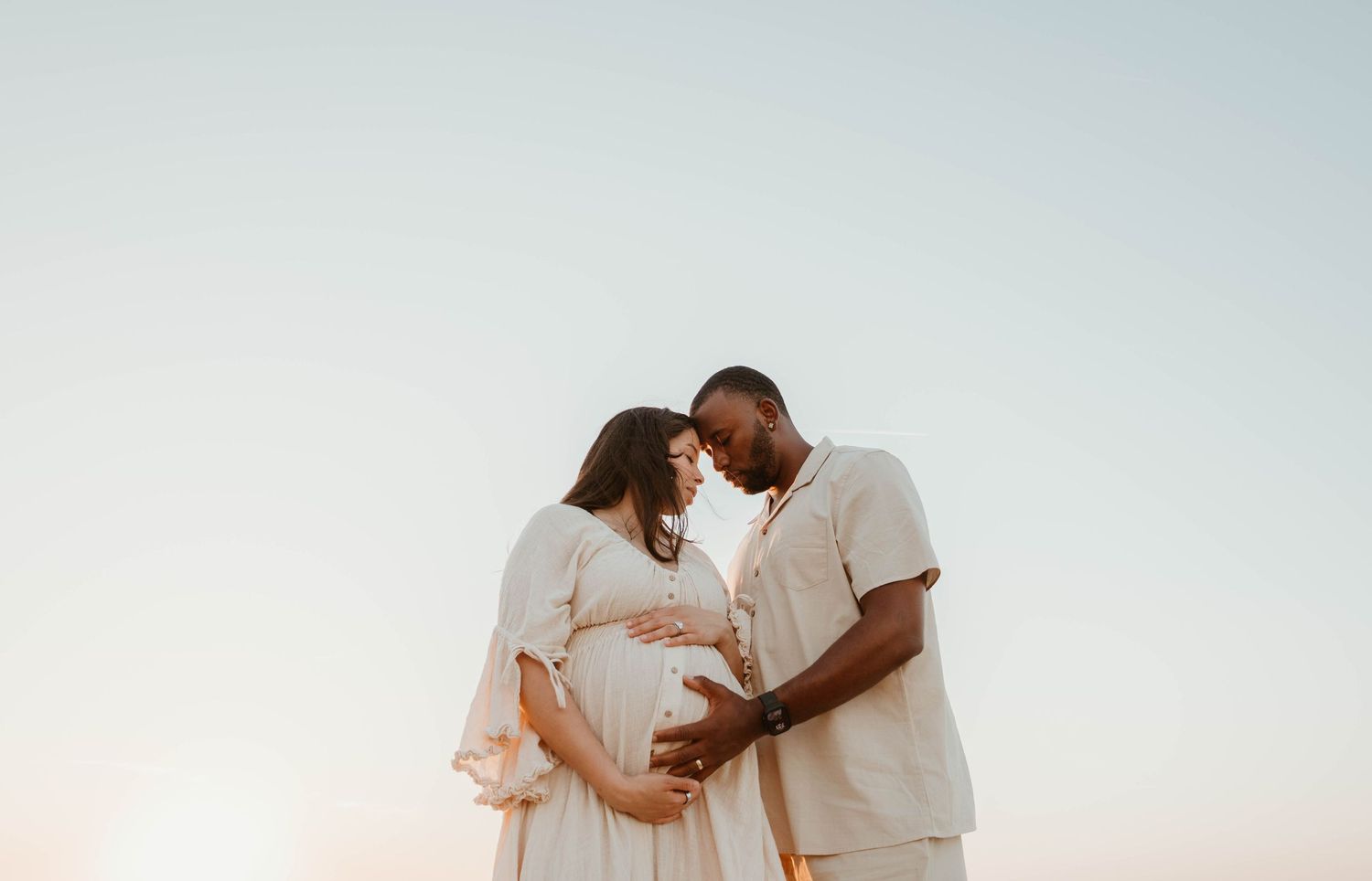 Pregnant couple tenderly touching foreheads outdoors at sunset, woman in white dress with hands on baby bump.