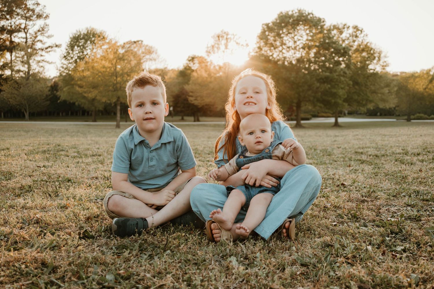 Three young siblings sit together on grass at golden hour, with sunlight glowing through trees in a park.