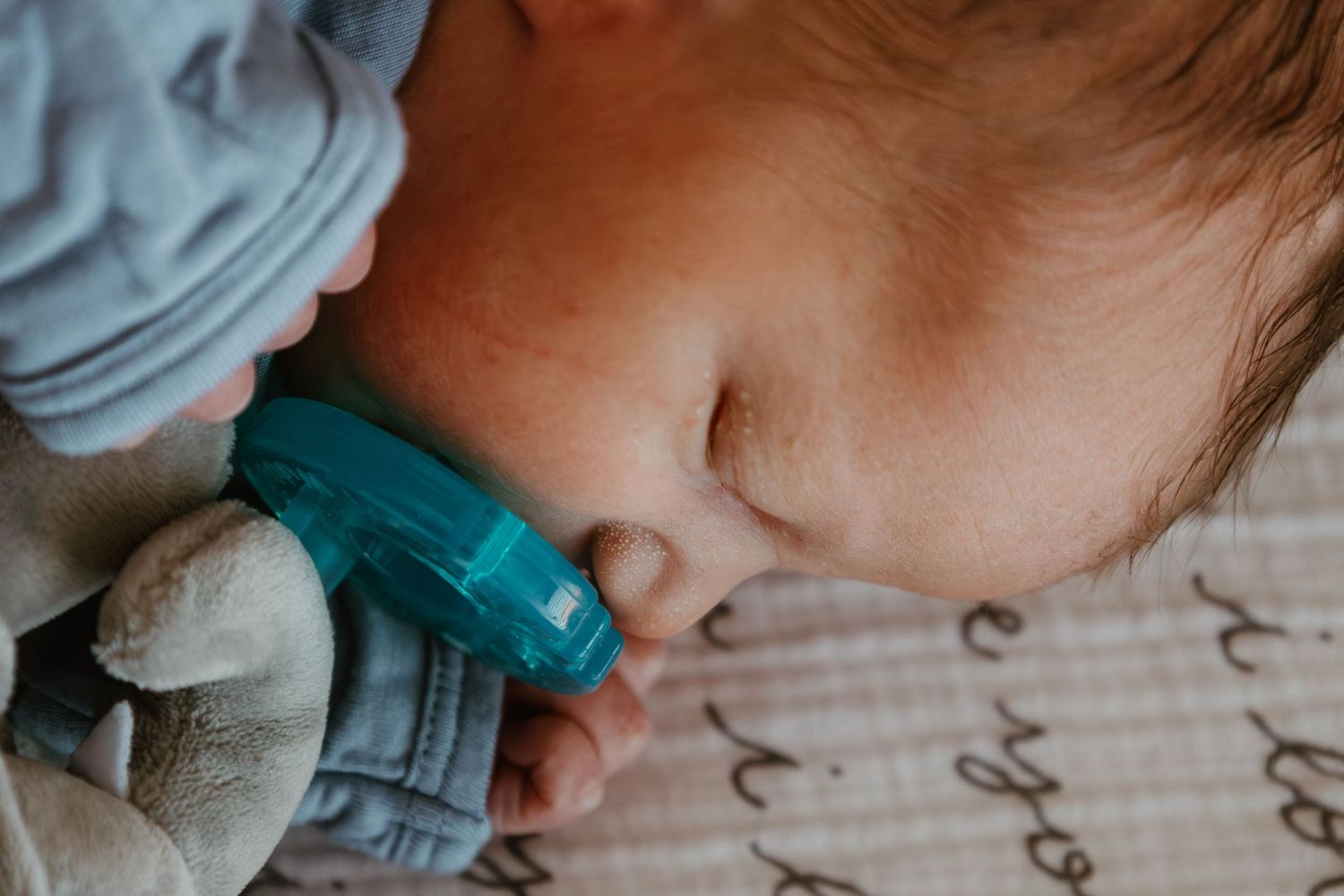 A sleeping baby wearing a teal pacifier and gray clothing rests peacefully on soft bedding.