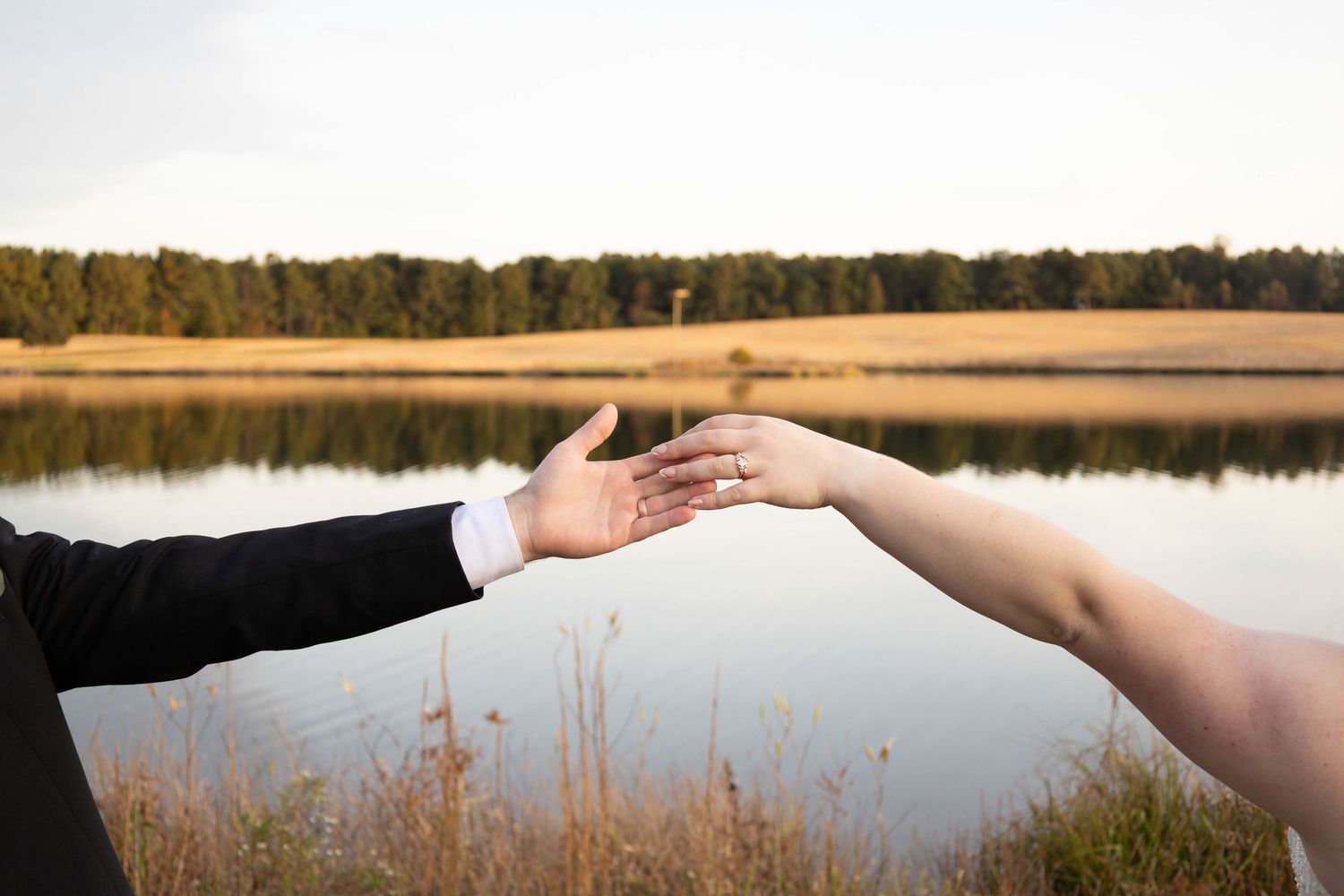 Hands reaching across water with wedding rings during sunset at lakeside ceremony.