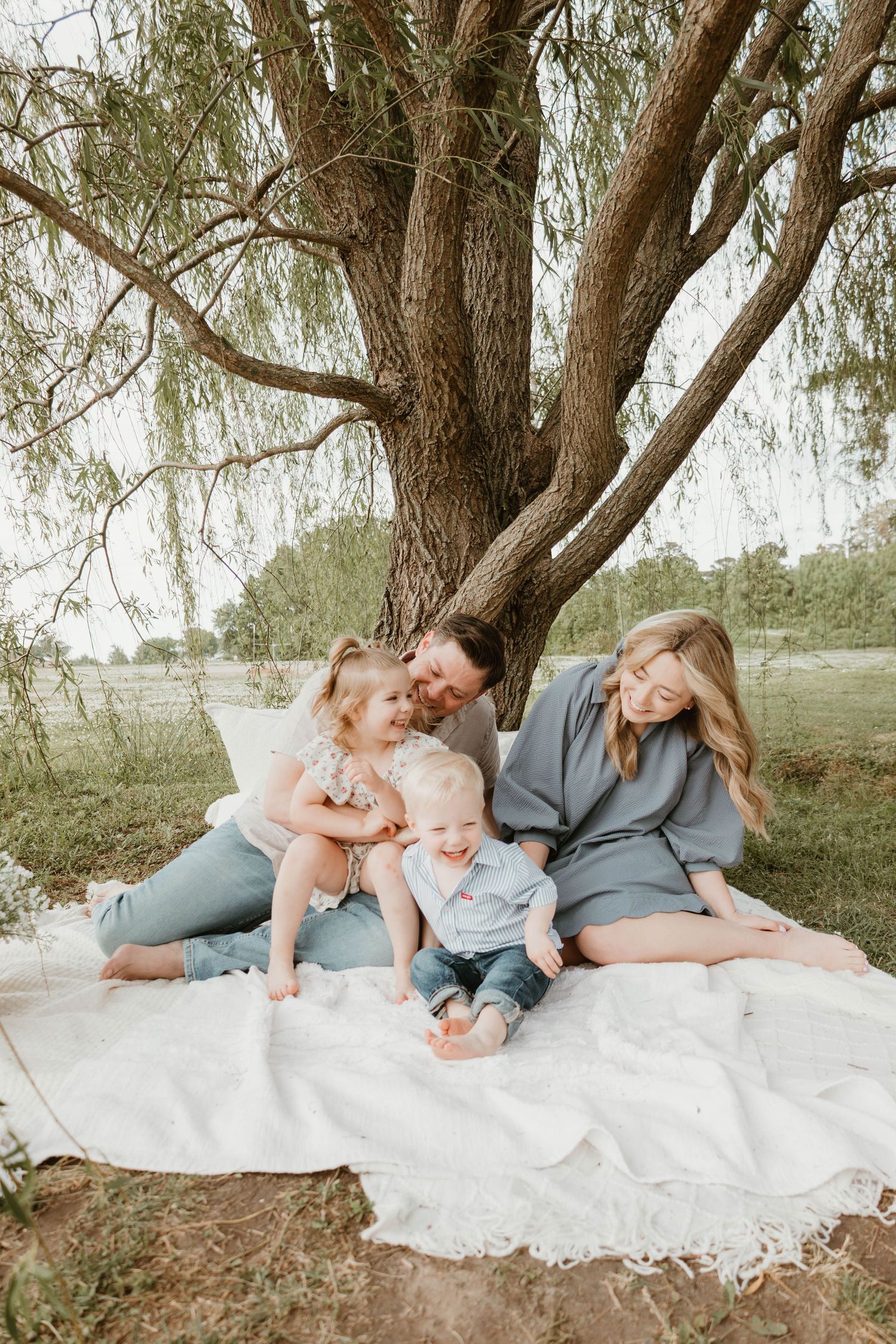 Family of four sitting on white blanket under willow tree, parents smiling at toddler and baby in outdoor portrait.