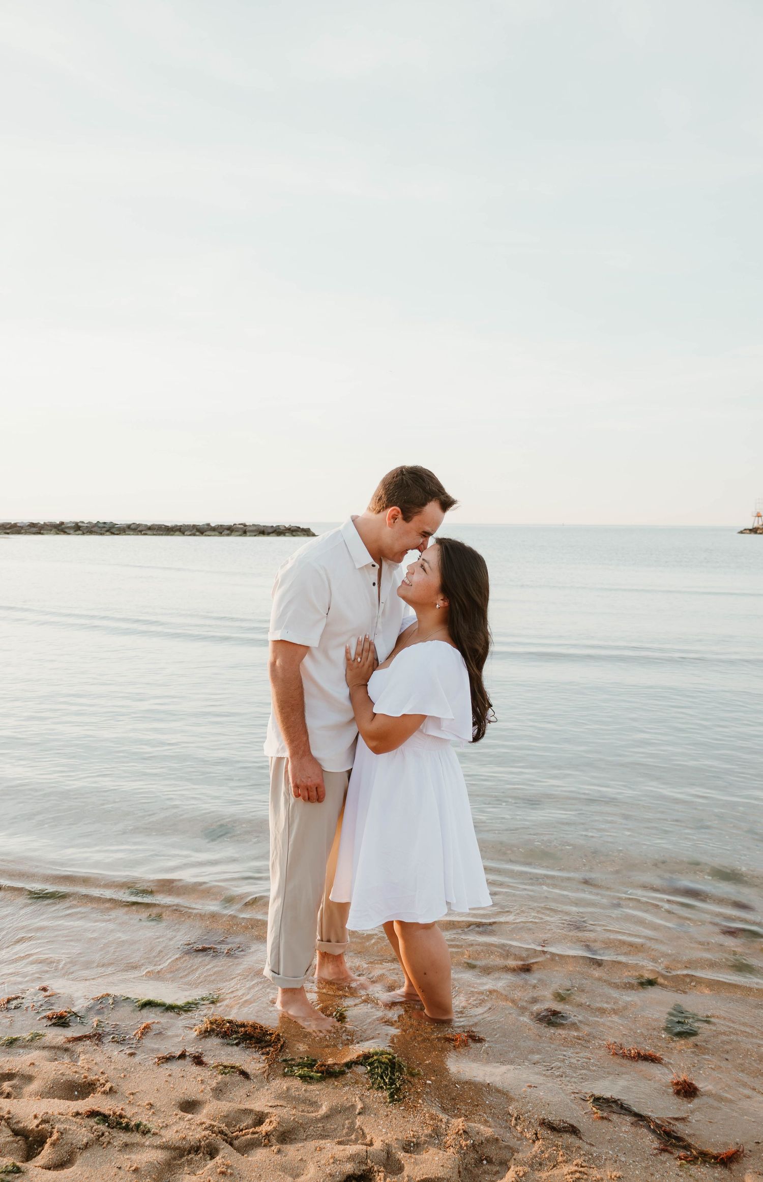 Couple sharing a romantic moment on a sandy beach, woman in white dress and man in casual attire, ocean backdrop.