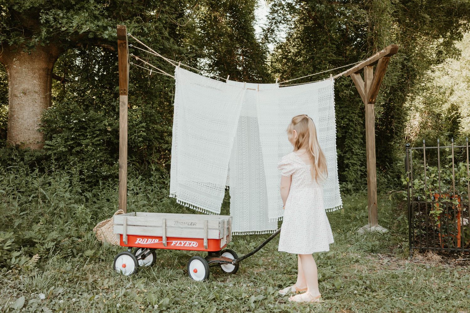 Young girl in white dress standing by a red Radio Flyer wagon near laundry hanging on a clothesline outdoors.