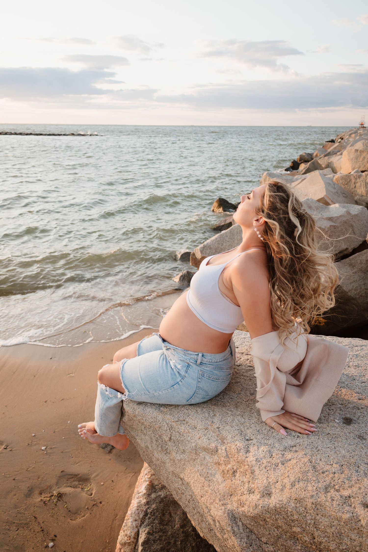 Woman in denim and white top relaxing on coastal rocks at sunset.