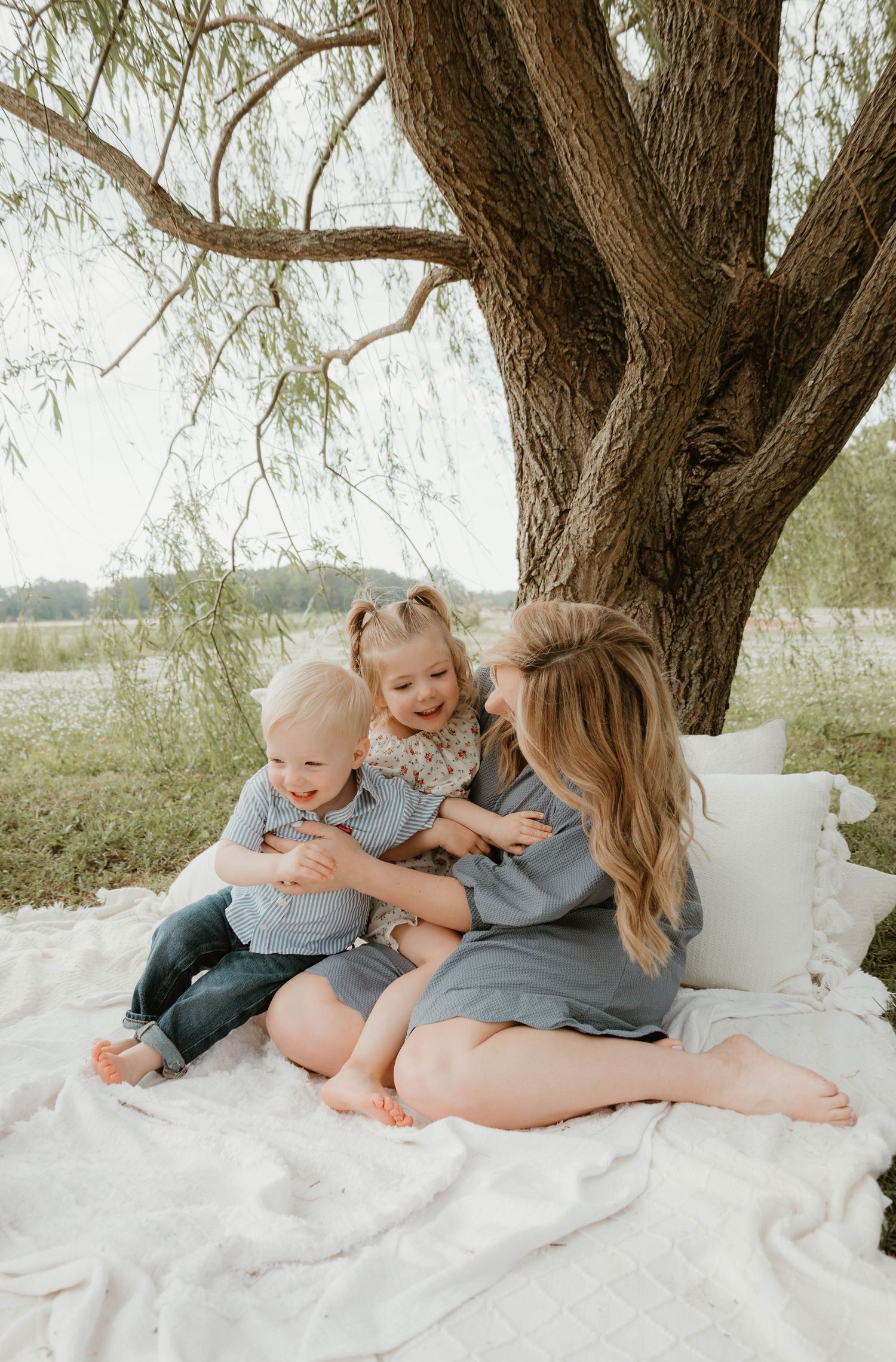 A mother sits on a white blanket under a large tree, laughing and cuddling her two young children outdoors.