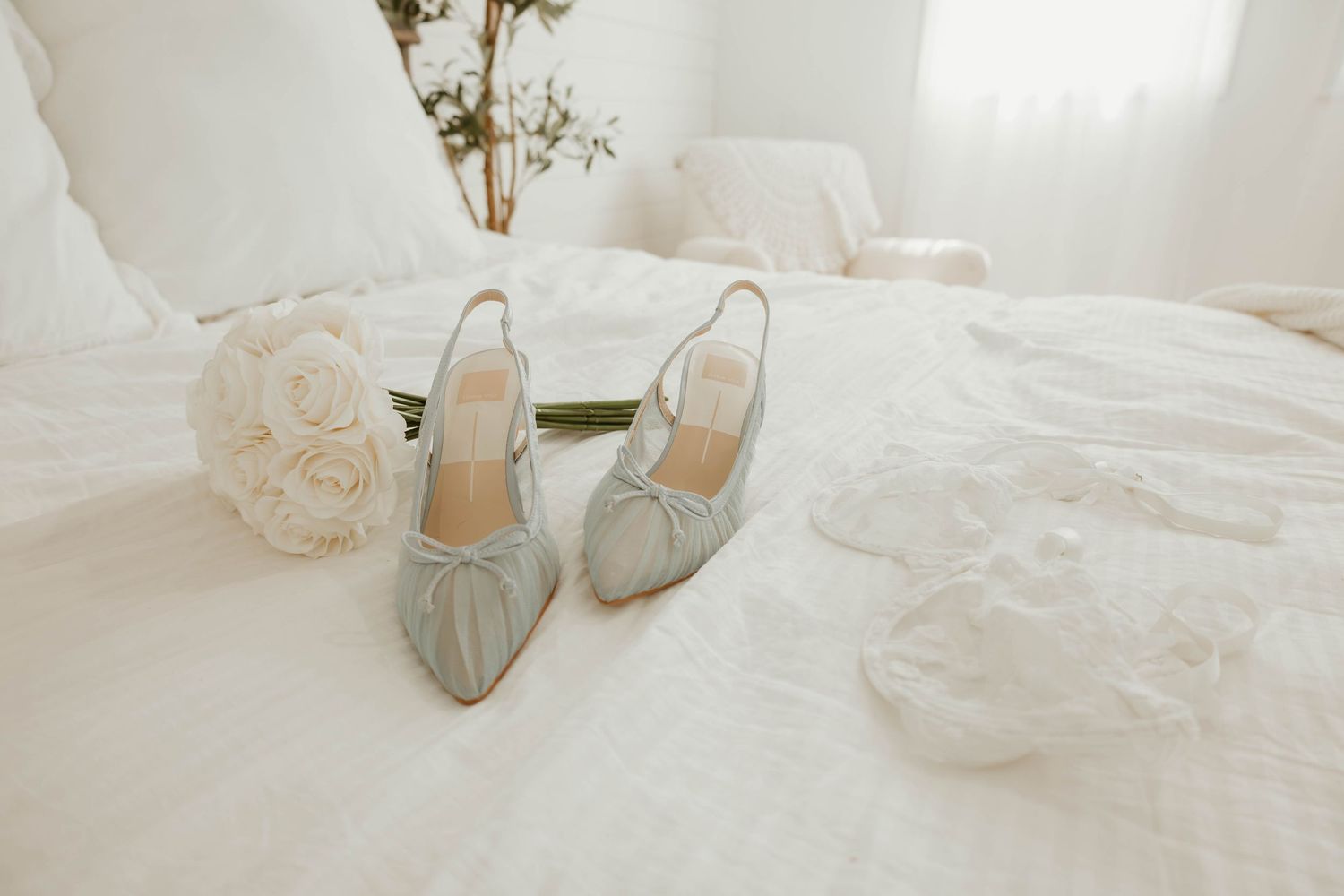 Elegant light blue slingback heels with bow detail beside a white rose bouquet on a white bed.