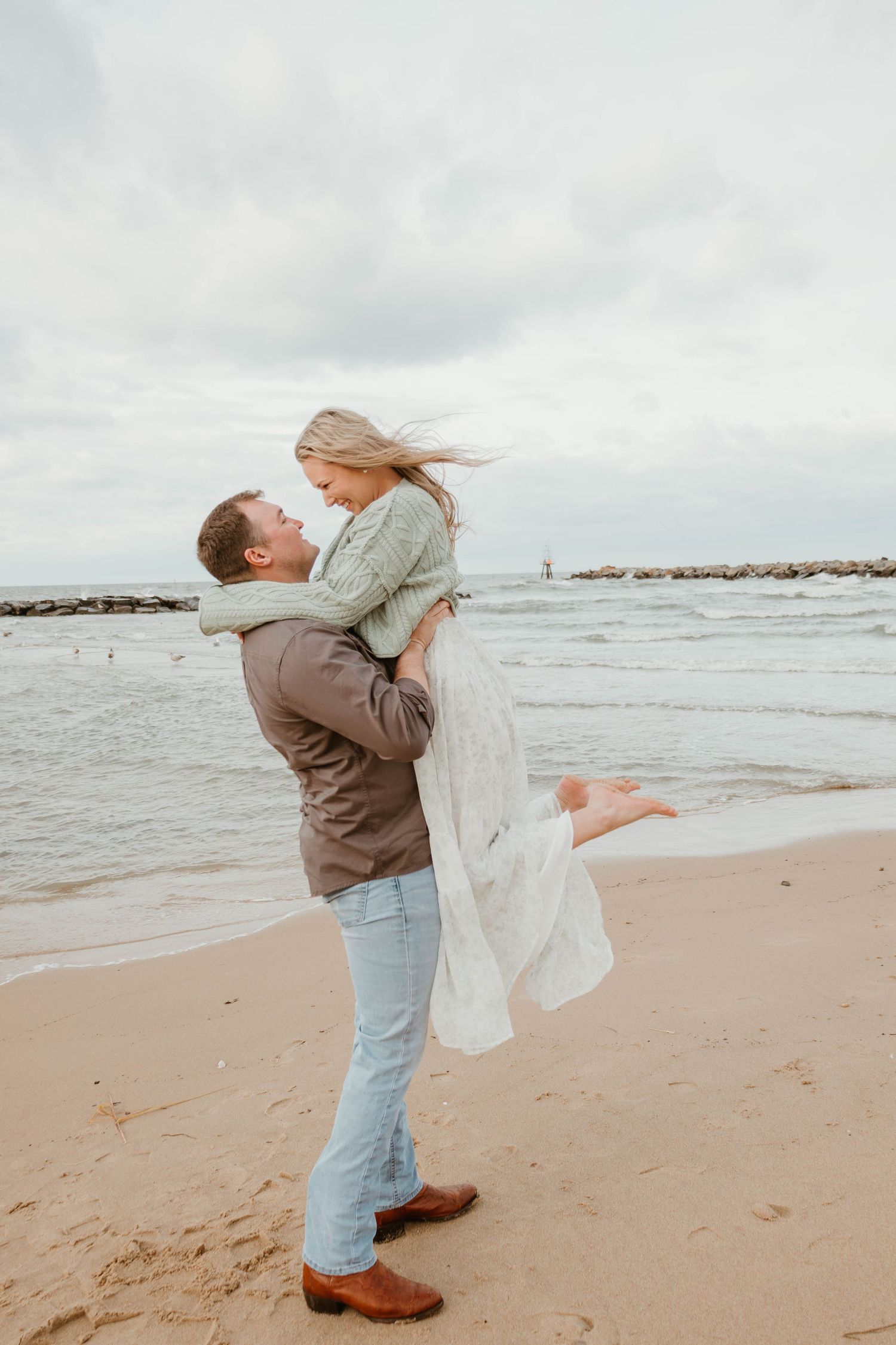 A couple embraces and spins joyfully on a cloudy beach with waves crashing in the background.
