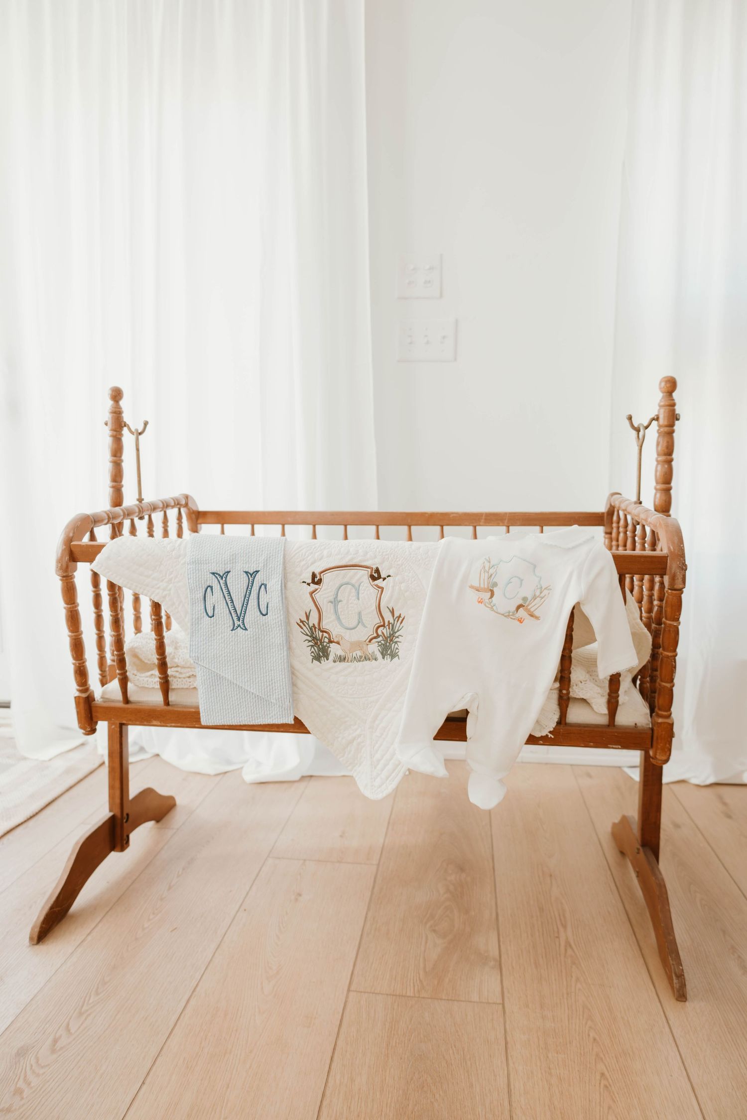 Vintage wooden rocking cradle with white linens displayed on light hardwood flooring against a white wall.