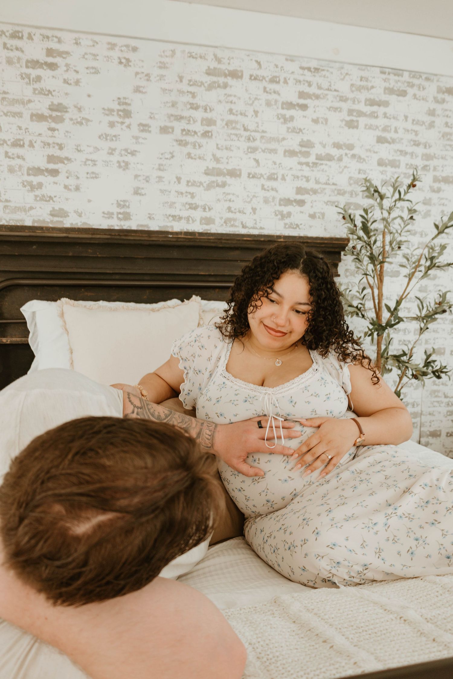 Pregnant woman in floral dress sitting on bed smiling at partner during intimate maternity photo session.