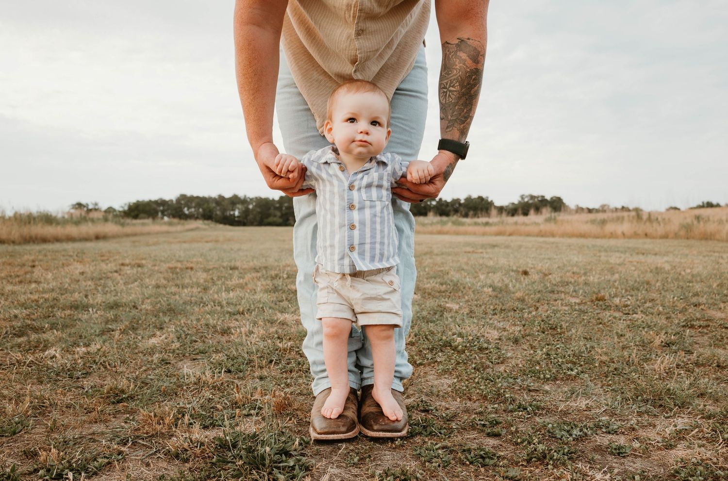 Adult helping a baby take first steps outdoors in a grassy field, tattooed hands guiding the toddler wearing a striped outfit.