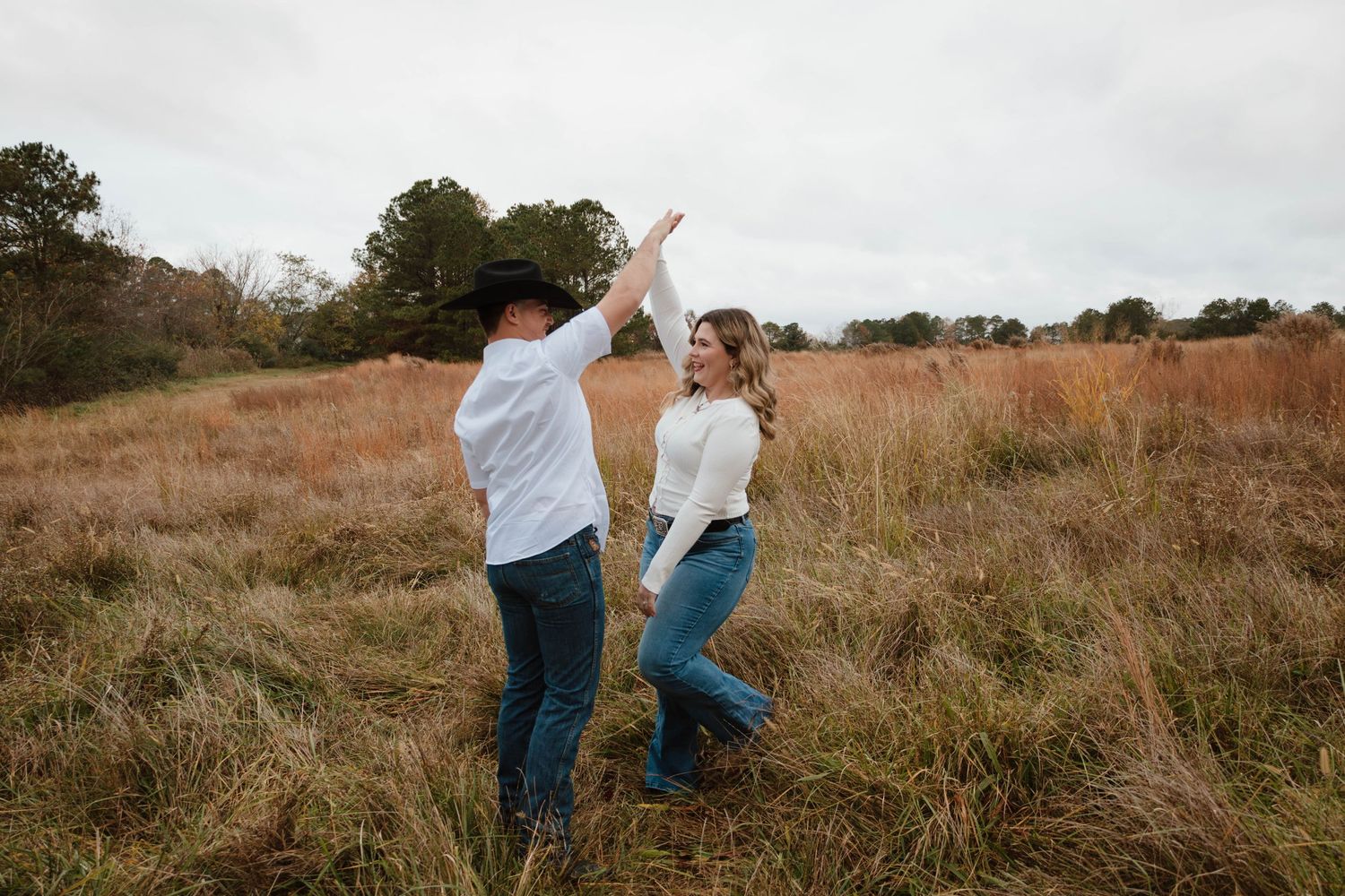Couple jumping and celebrating together in tall grass field during overcast day.
