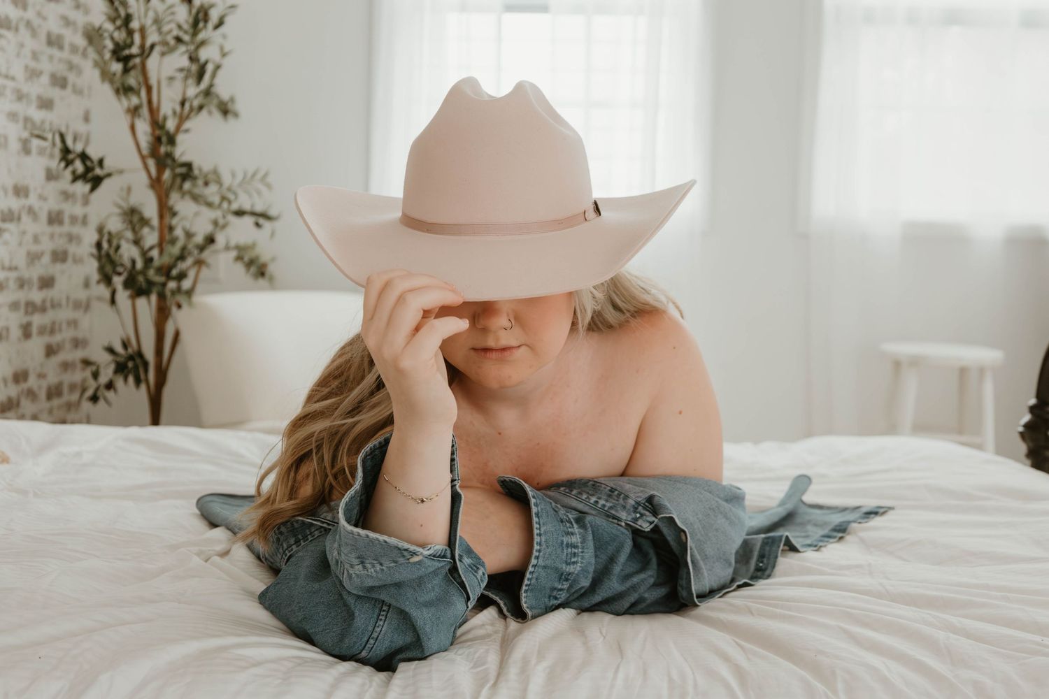 Woman in pink cowboy hat and denim jacket lying on white bed in bright room.