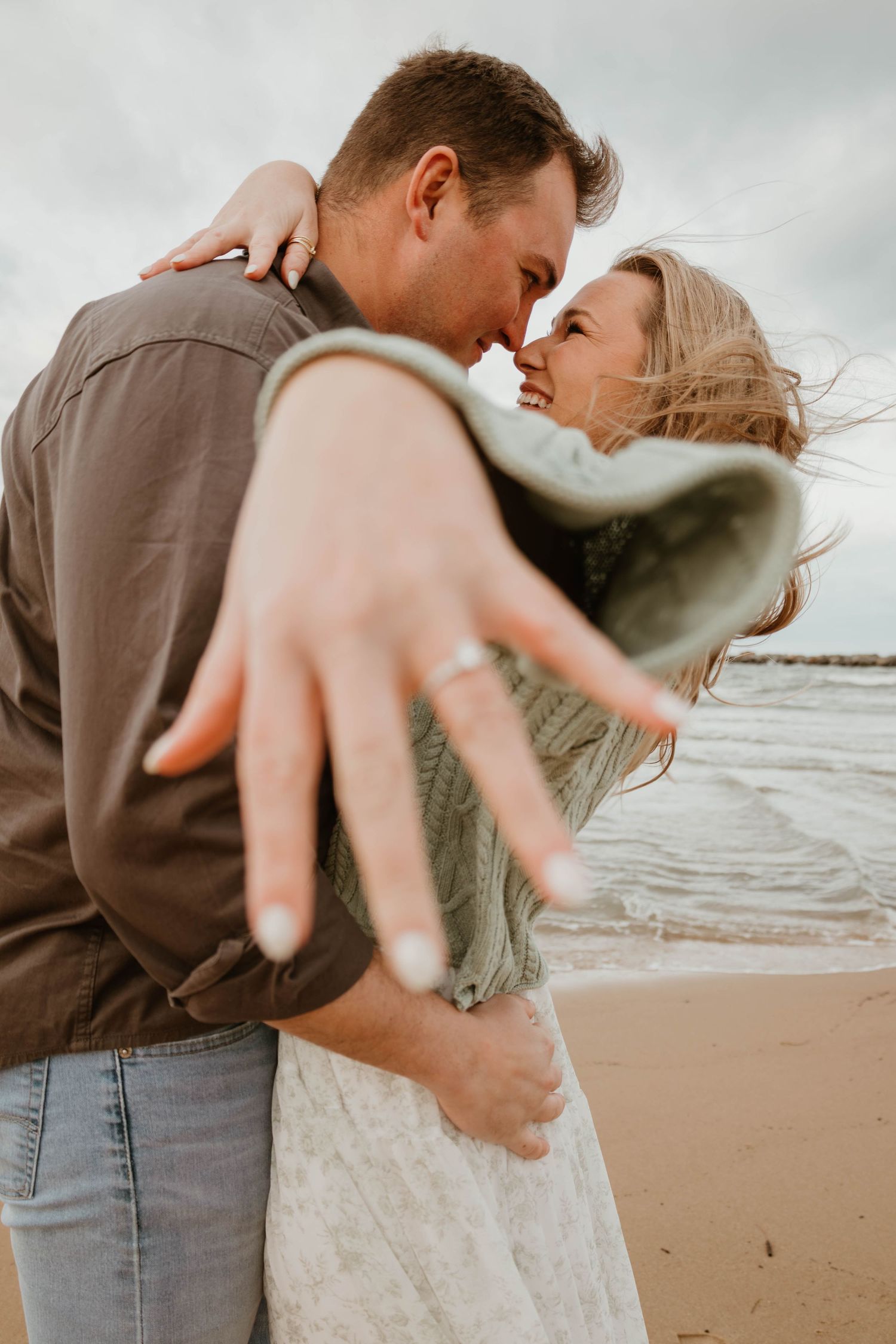 A romantic couple embraces on a cloudy beach, with wind blowing through their hair as they share an intimate moment.