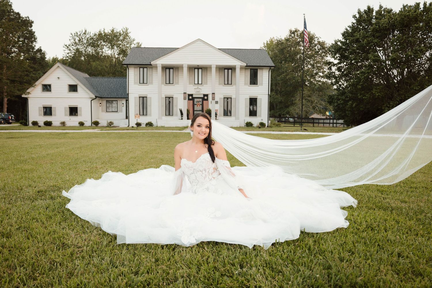 A flowing white wedding dress train spreads across a grassy field near a white mansion.