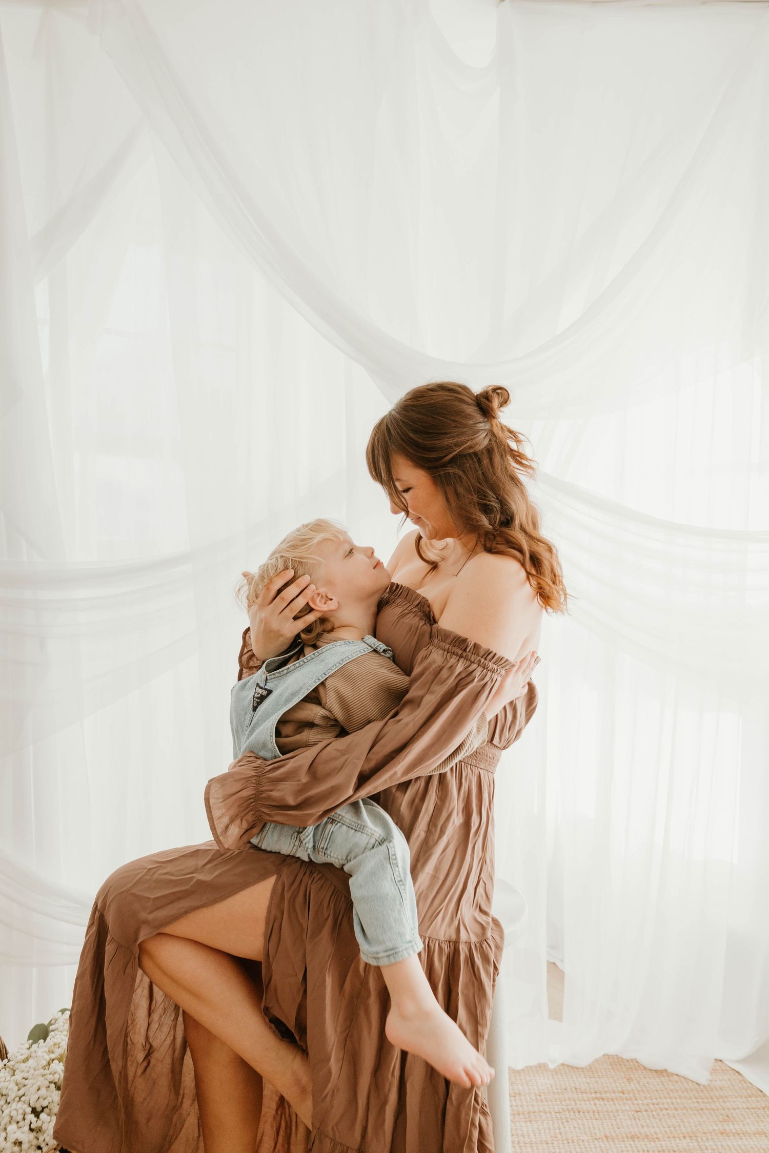 A tender moment captured in soft light as a mother and child embrace in matching neutral-toned outfits against white fabric.