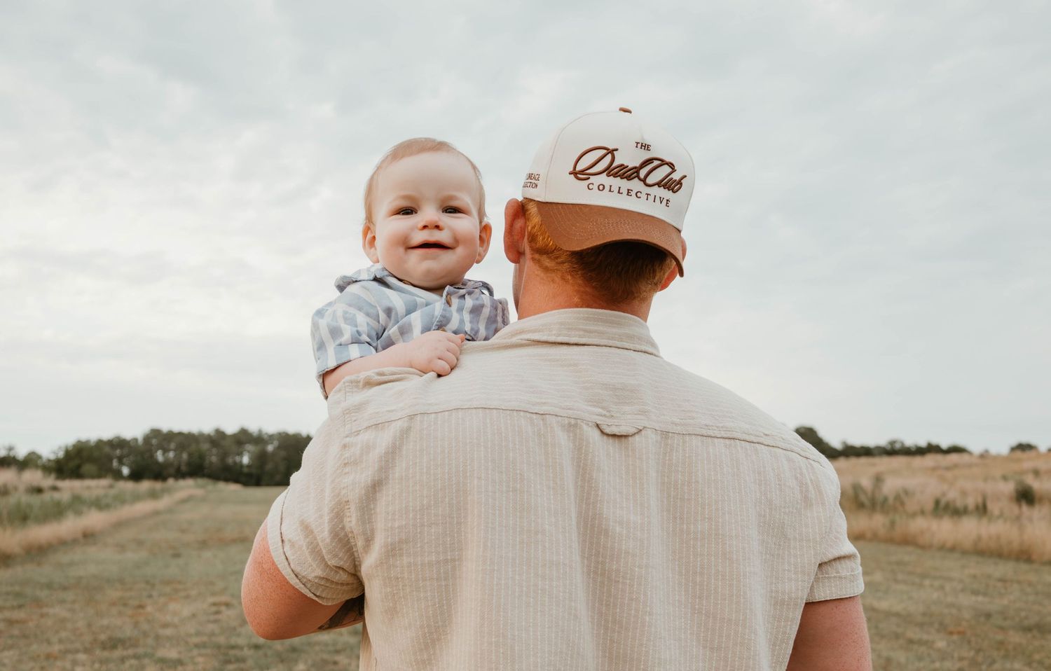 A smiling baby held on the shoulder of a man in a white cap and shirt in a rural field setting.