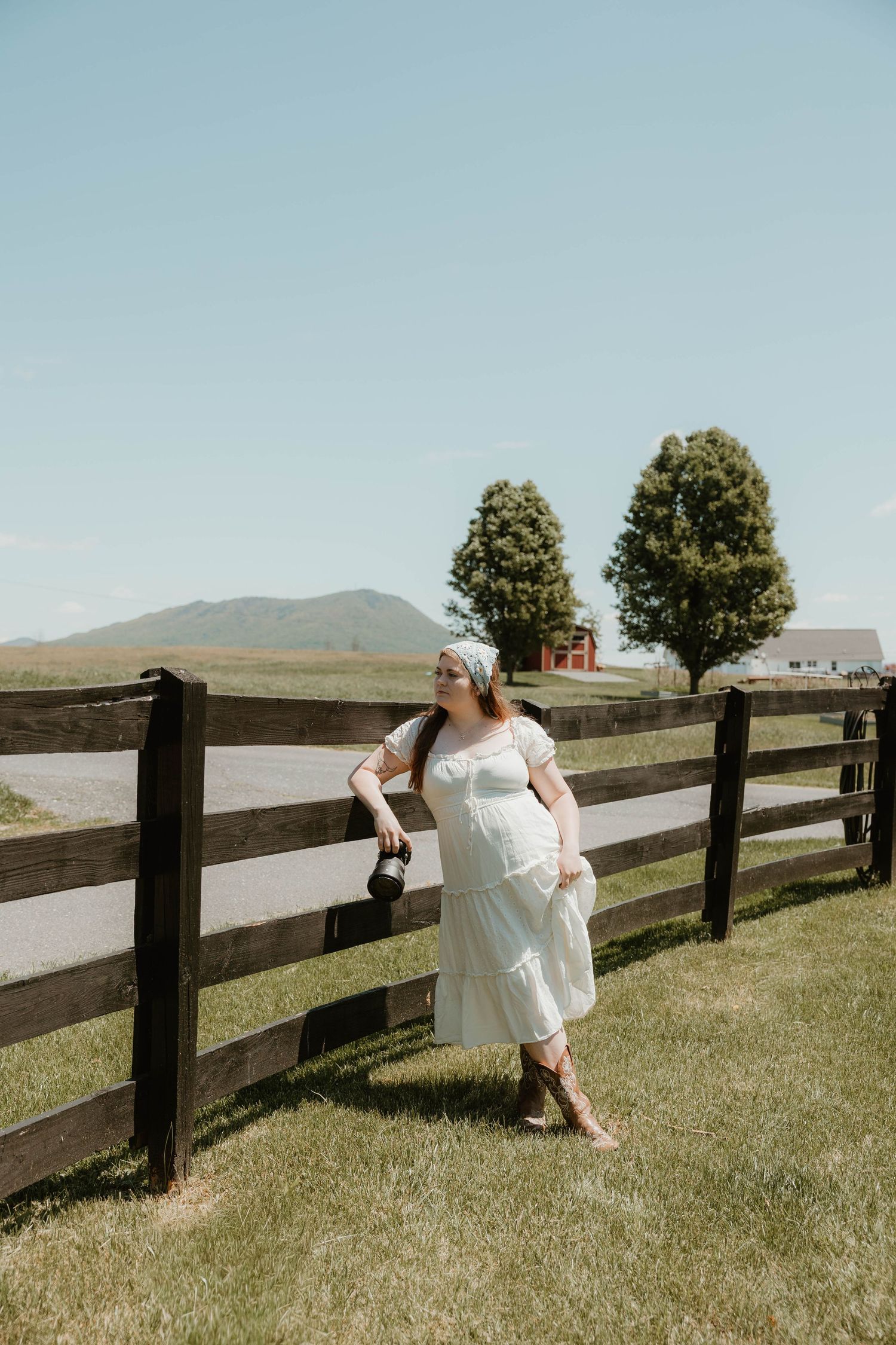 Woman in white dress leaning on wooden fence in rural countryside with mountain and trees in background.