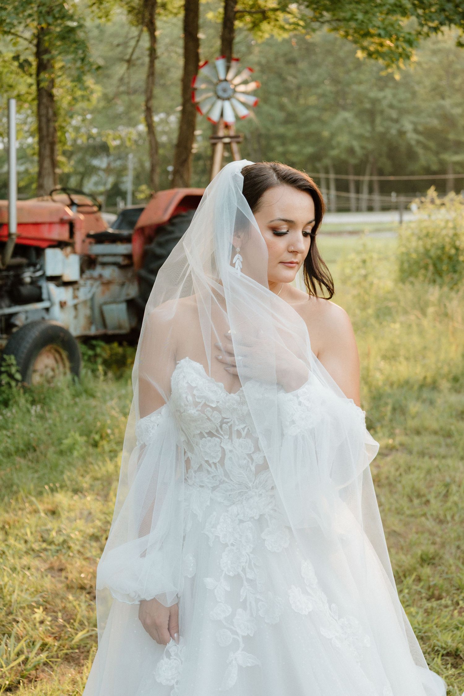 A bride in an ornate lace wedding dress poses in front of a vintage tractor and windmill.