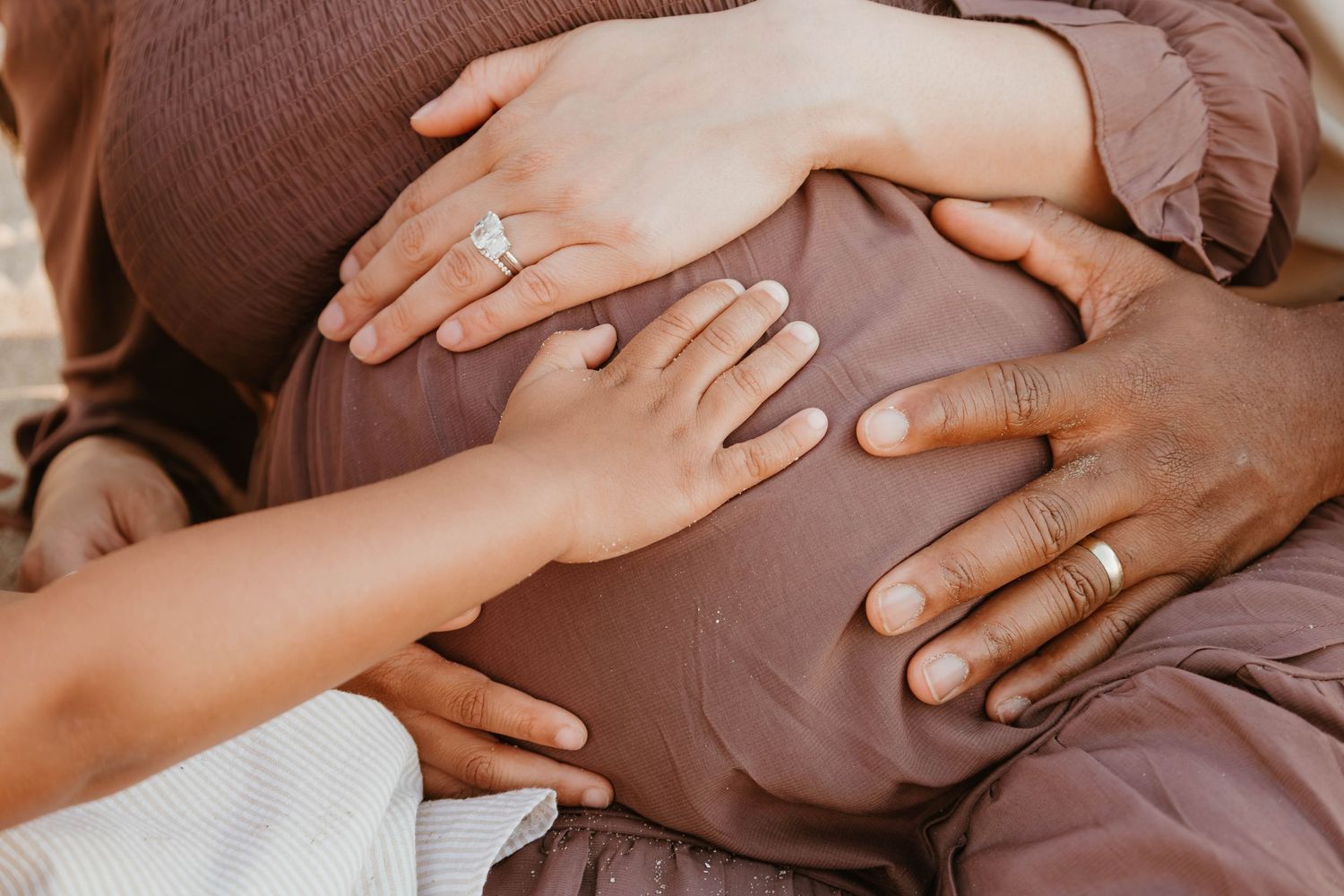 Pregnant woman in mauve dress with multiple hands gently cradling her baby bump, wearing an engagement ring.