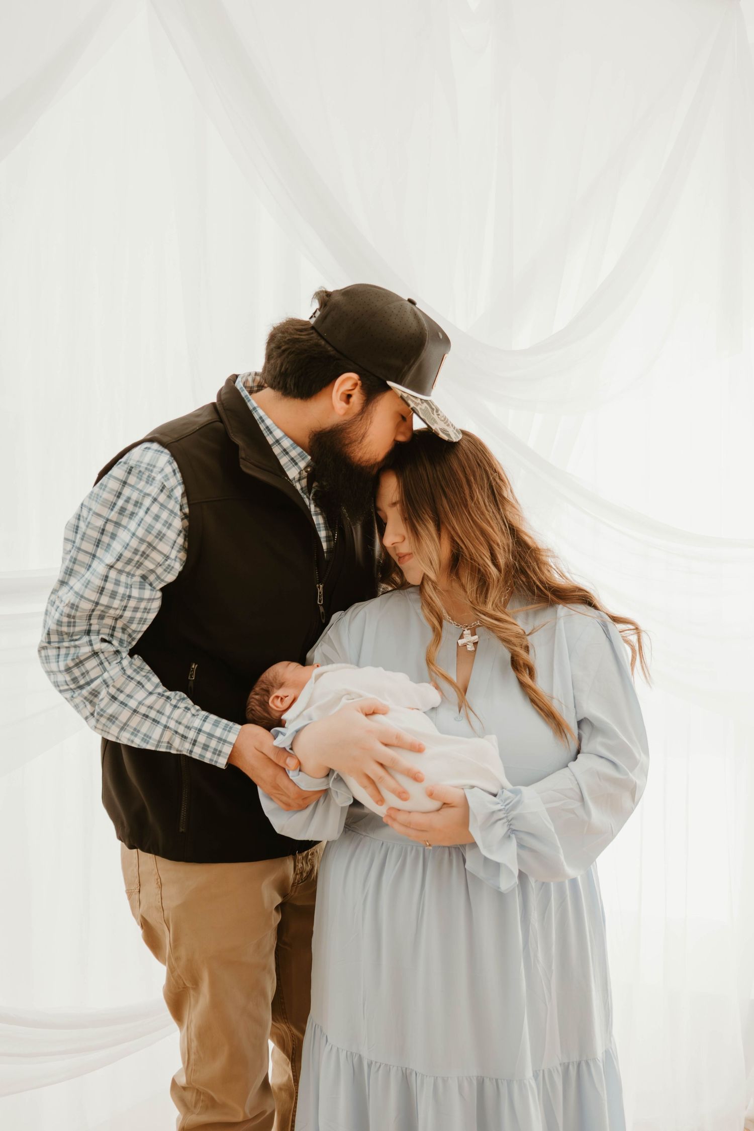 Parents in casual clothing tenderly embracing their newborn baby against white backdrop.