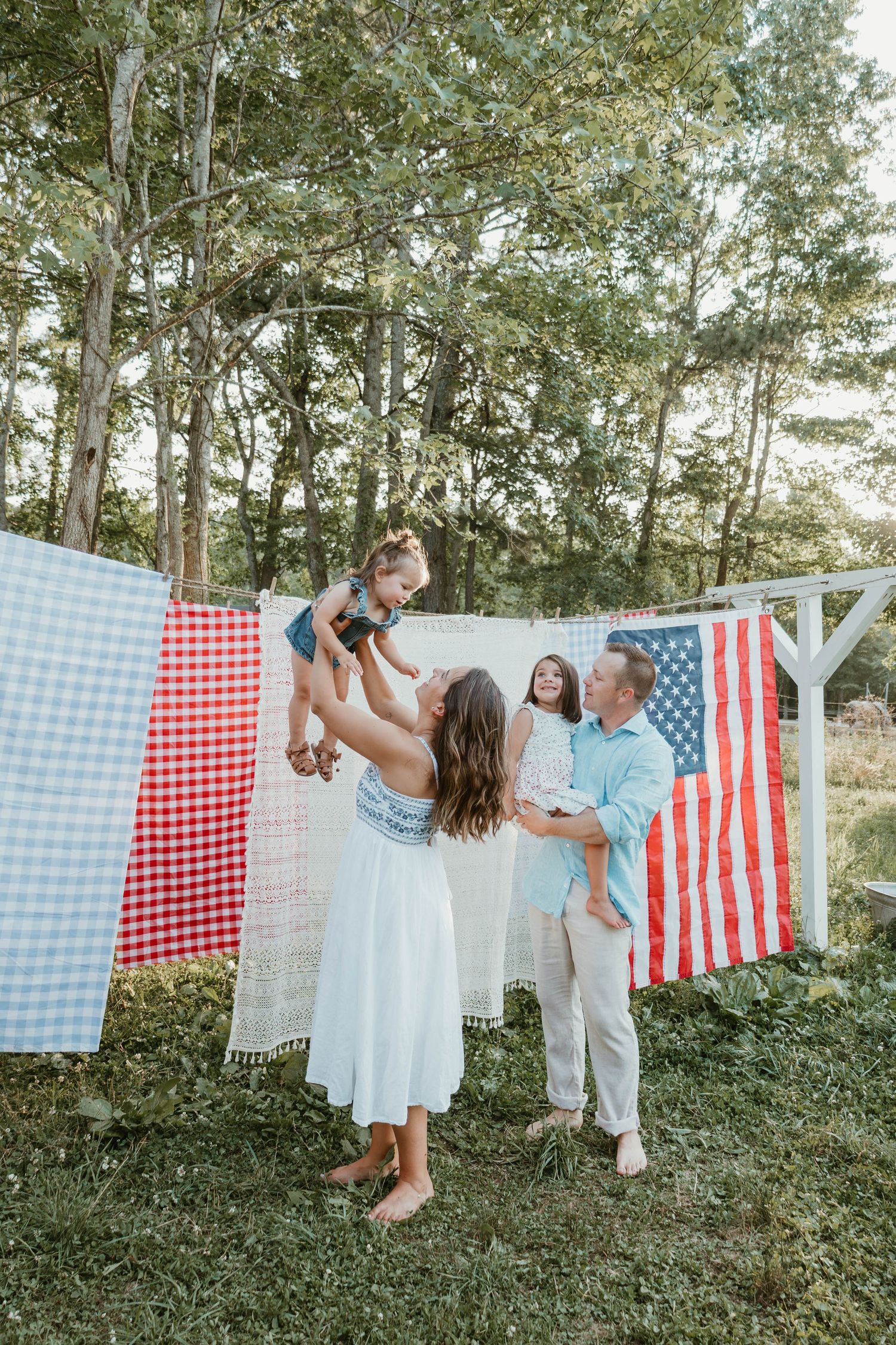 Family celebrating Fourth of July outdoors, lifting children near American flag and colorful quilts hung on a clothesline.