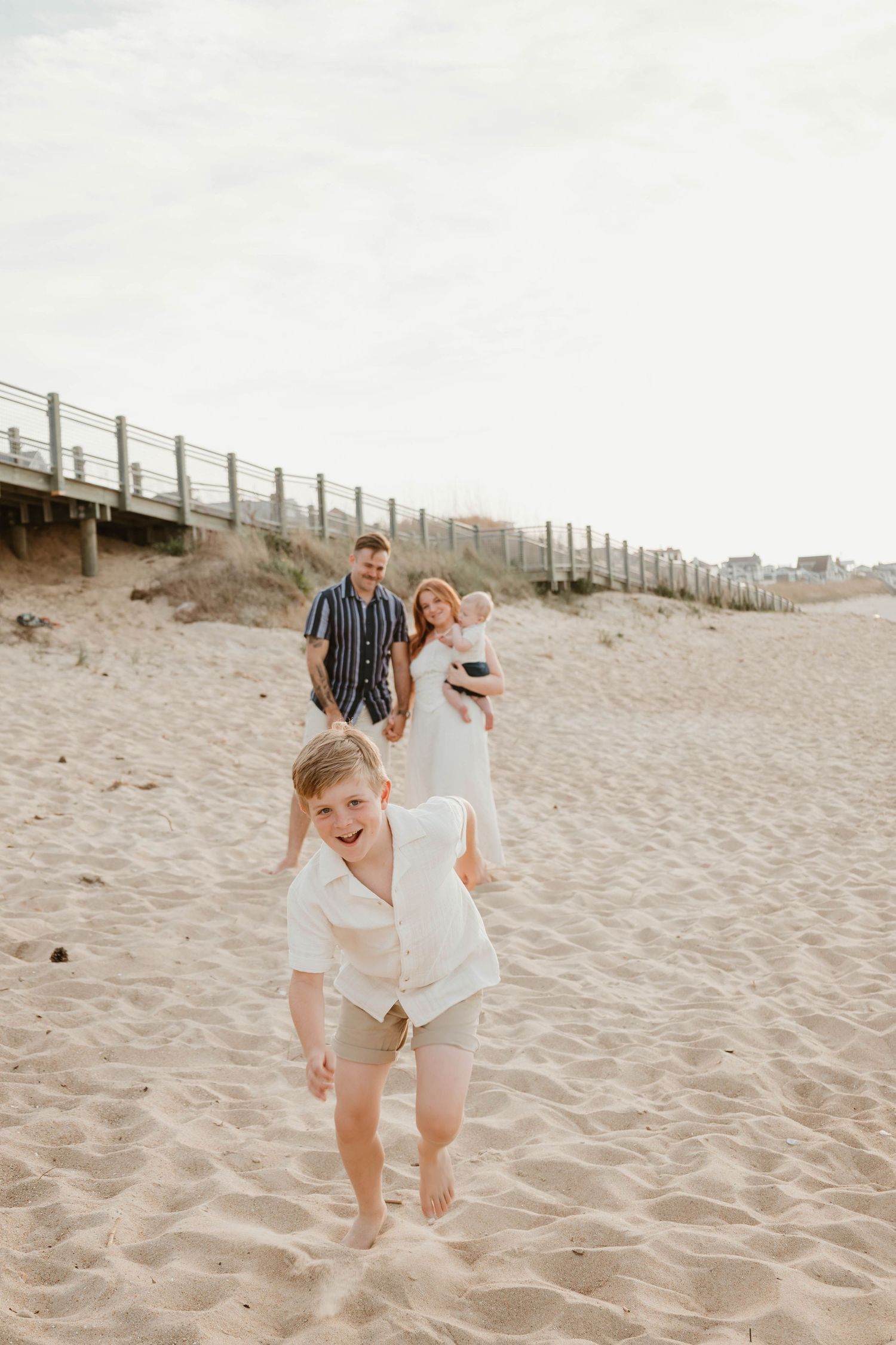 A joyful family of four enjoys a sunny beach day, with two children running and parents holding a baby on sandy shores.
