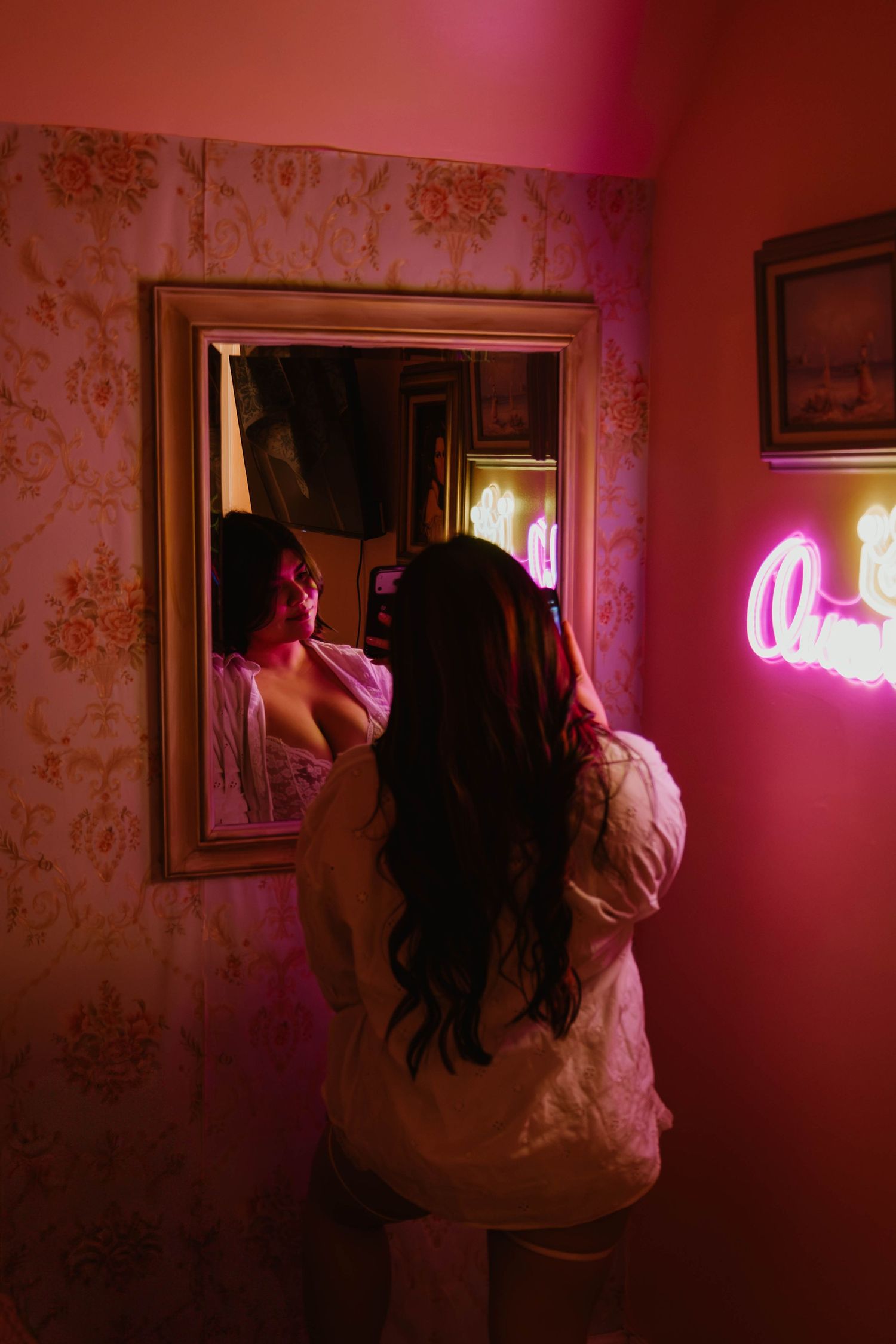 Woman with long dark hair in white robe gazes into ornate mirror beside pink neon sign in vintage floral wallpaper room.