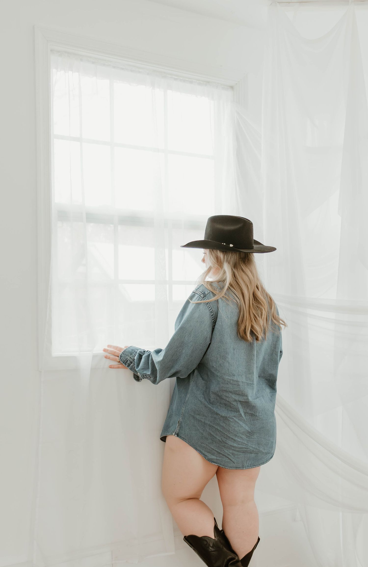 Woman in black hat and denim shirt standing by window in white room with cowboy boots.