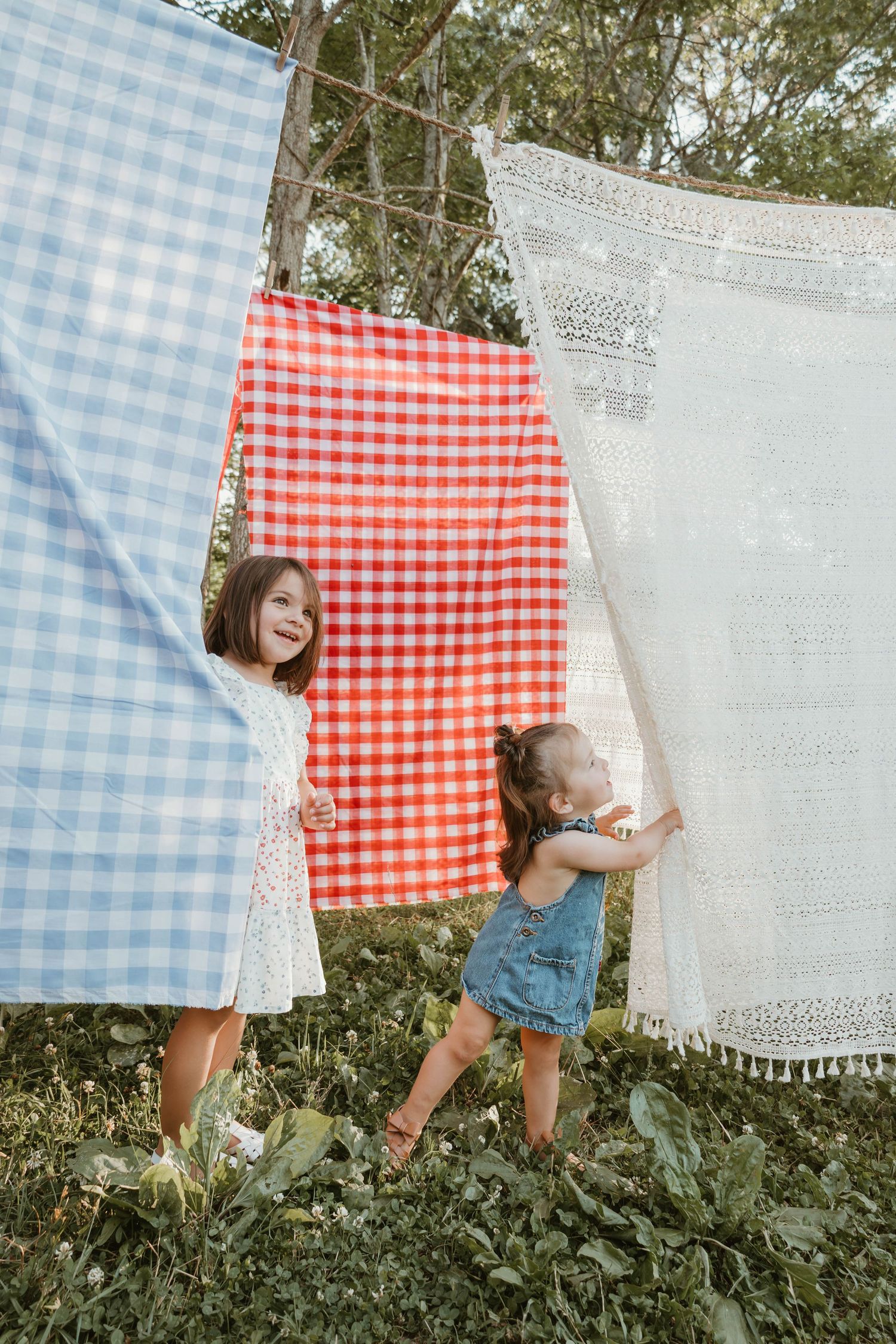 Two young girls playing joyfully among colorful gingham and lace tablecloths hanging on a clothesline outdoors.