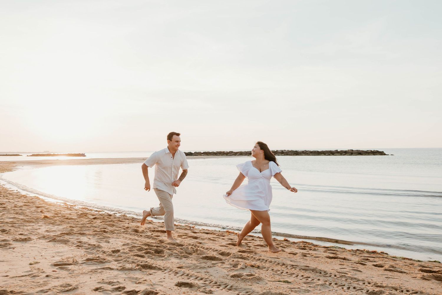 Couple running and laughing together on a sandy beach at sunset, wearing white outfits with calm ocean waters behind them.