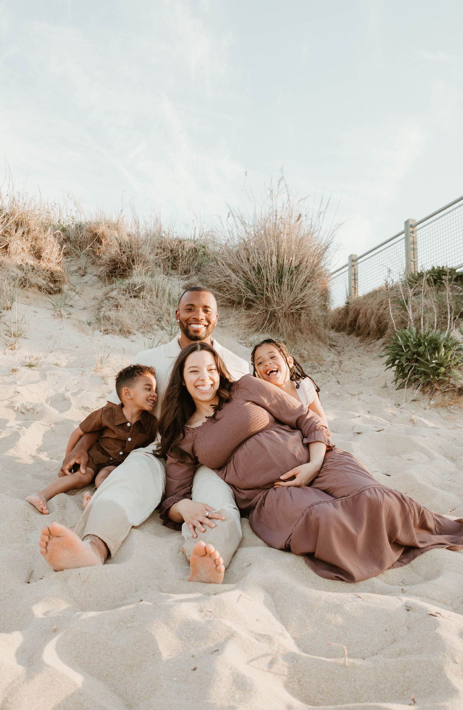 Happy family of four sitting on sandy beach dunes, smiling together during a maternity photo session on a sunny day.