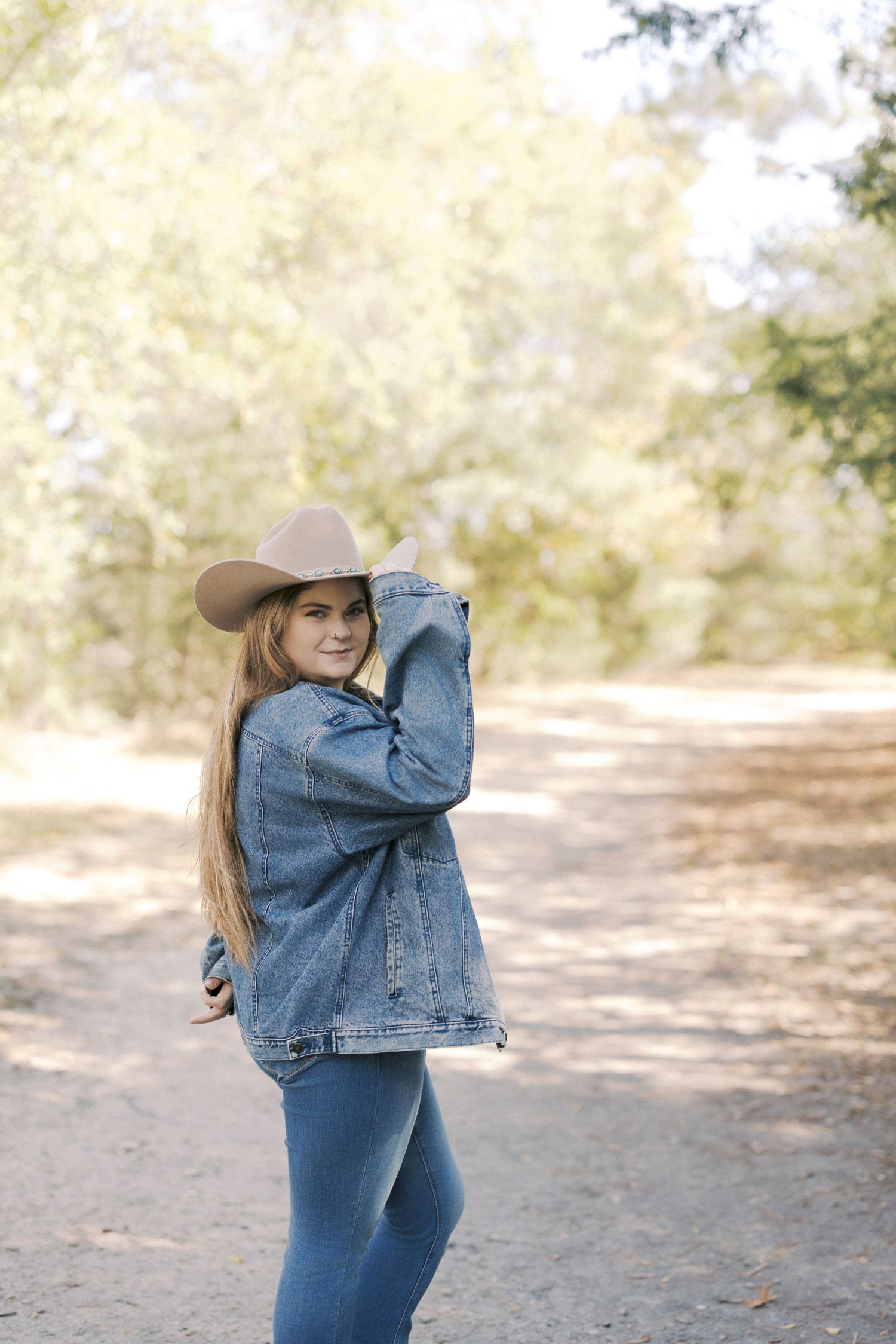 A person in a denim jacket and cowboy hat walks along a dirt path through a sun-dappled wooded area.