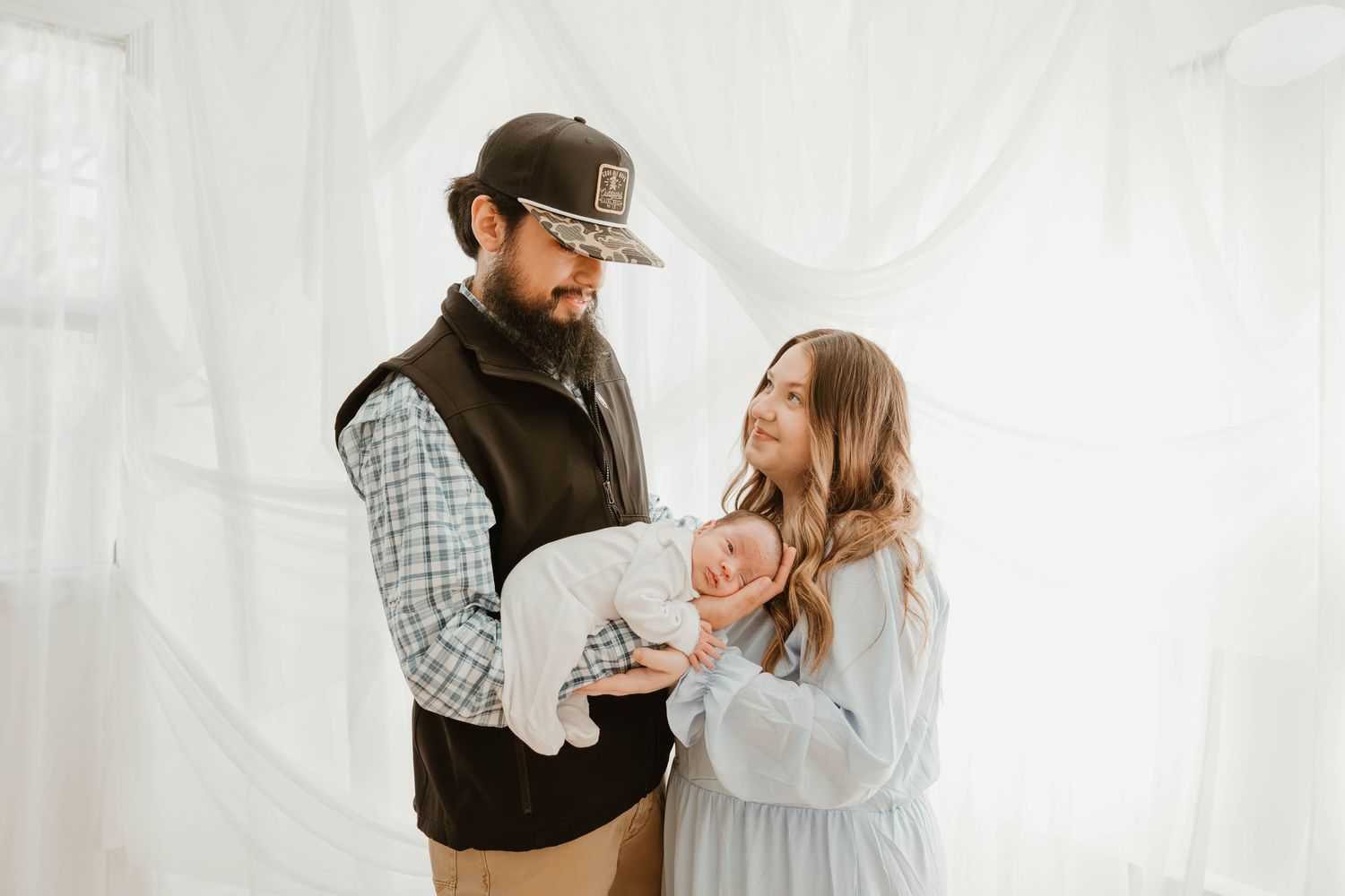 Parents in casual winter attire share a tender moment with their newborn baby against a white backdrop.