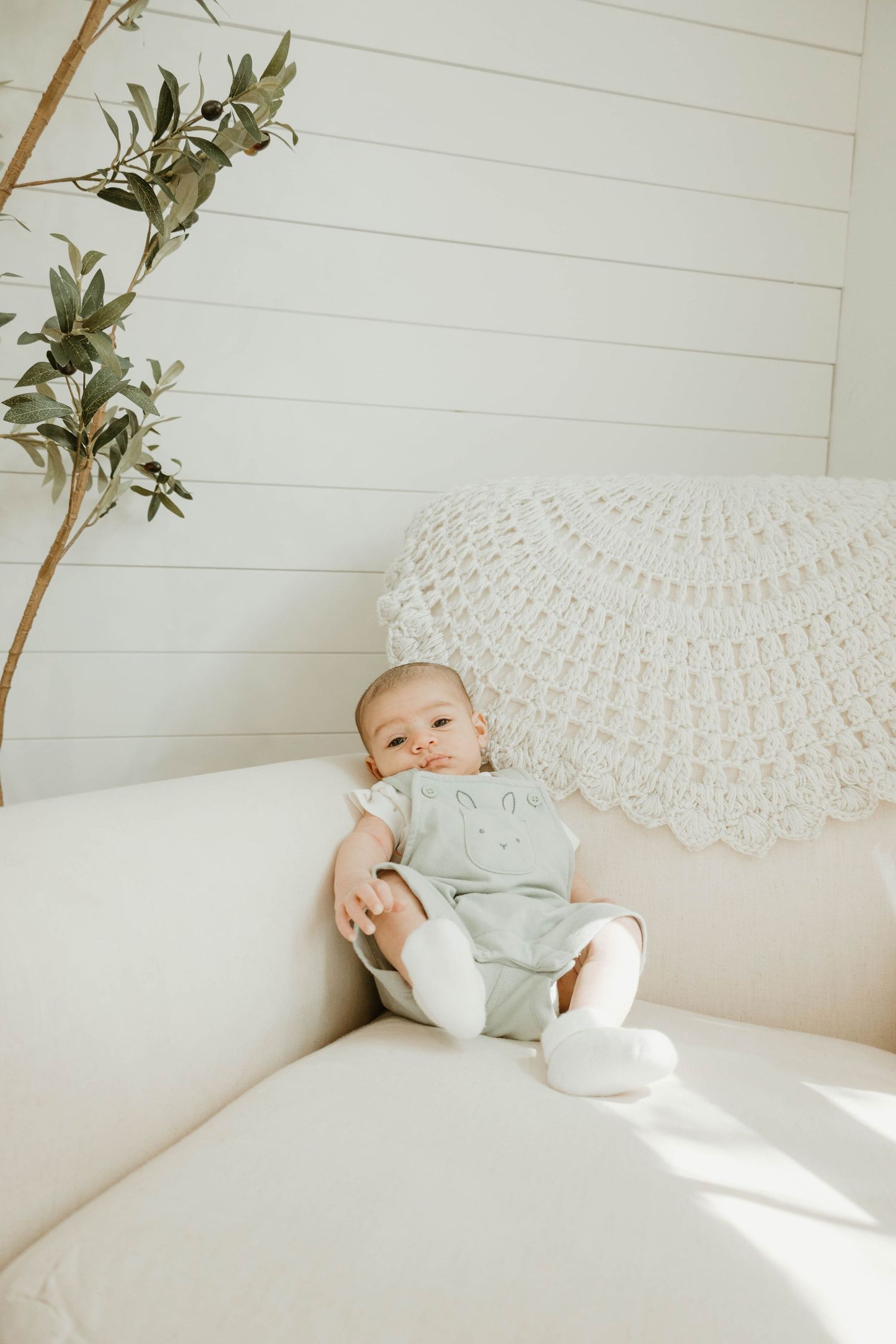 Adorable baby in sage green romper sitting on a cream sofa with crochet pillow and greenery in bright nursery.
