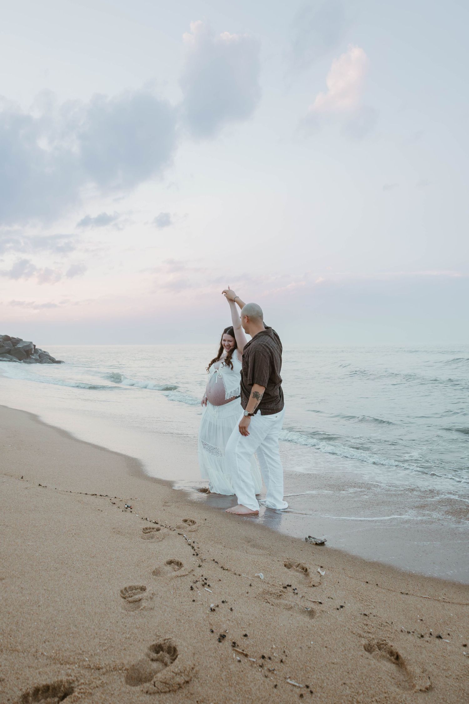 Couple dancing on beach at sunset, pregnant woman in white dress, footprints in sand, ocean waves in background.