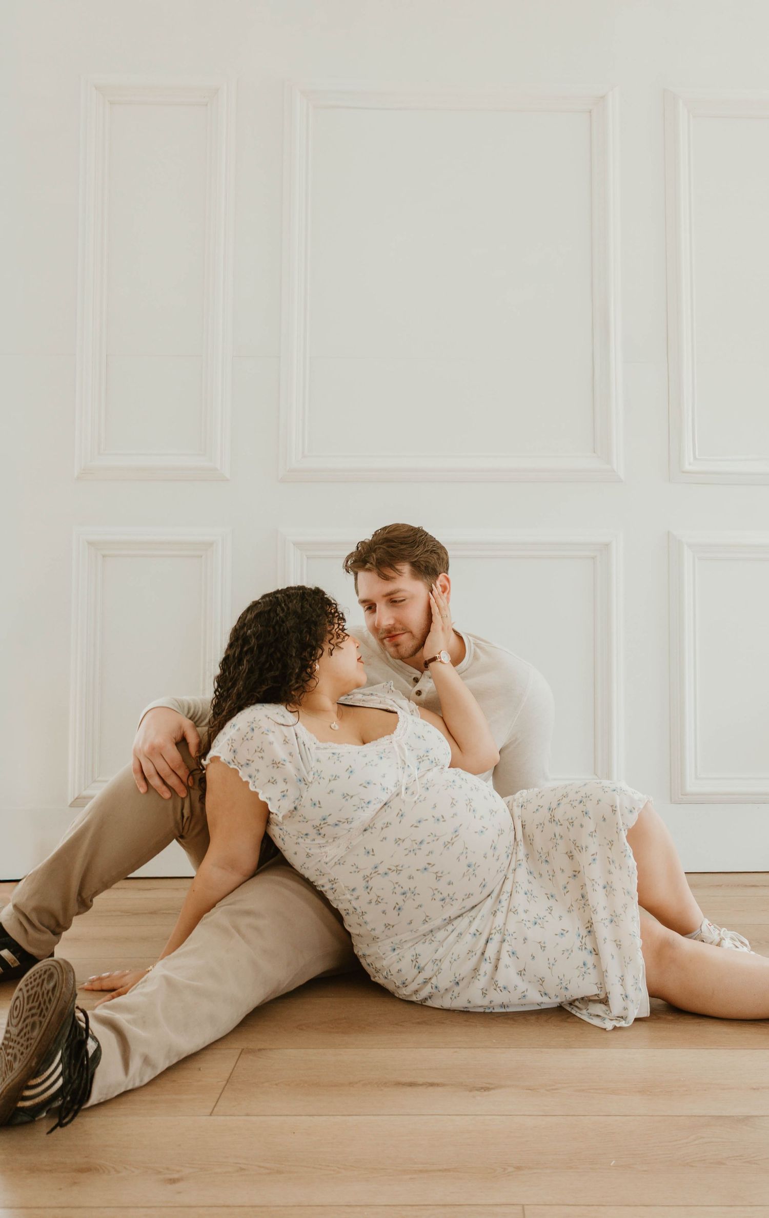 A couple sits together on the floor, embracing and looking affectionately at each other.