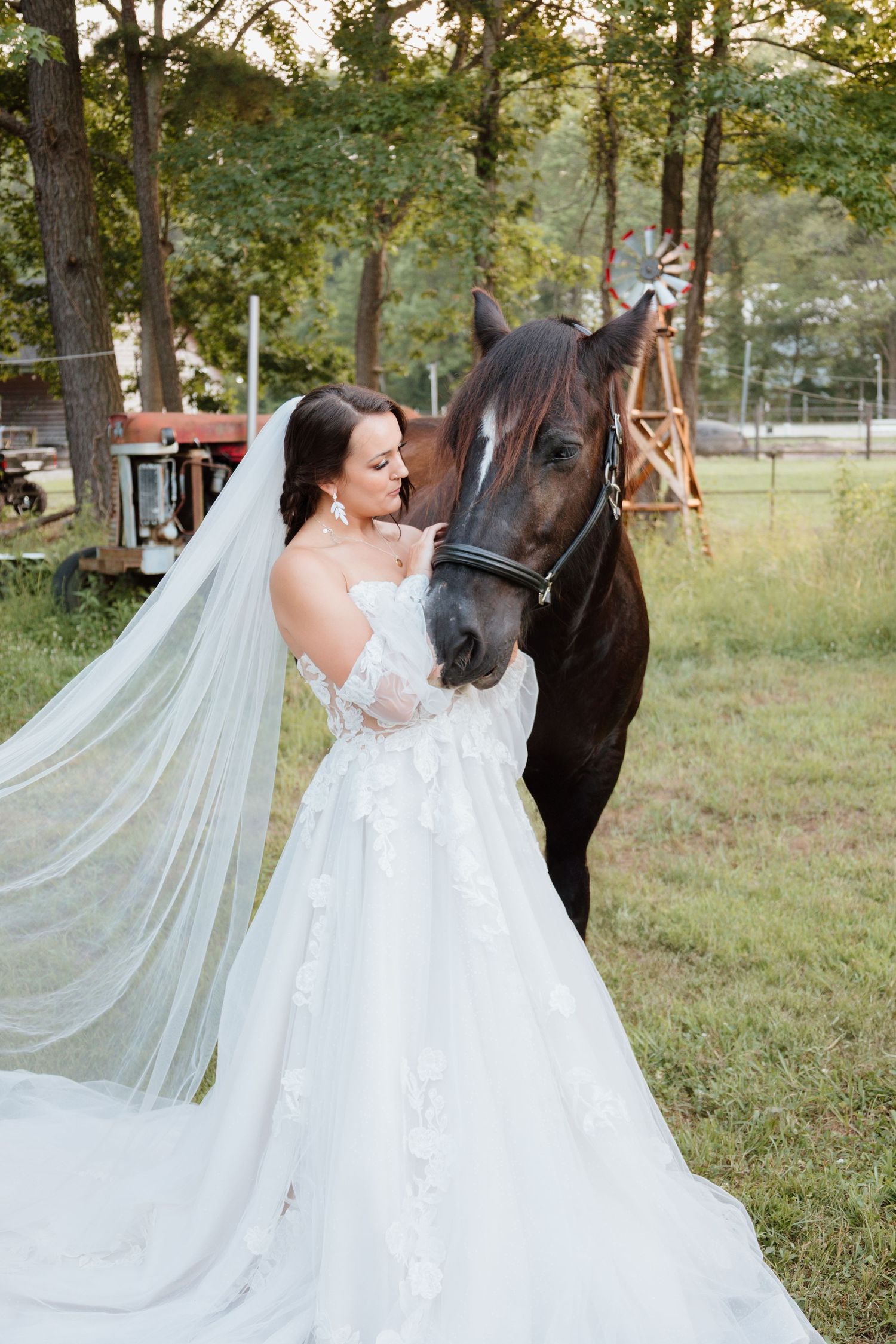 A bride in a flowing white gown stands with a dark horse in a rustic outdoor setting.