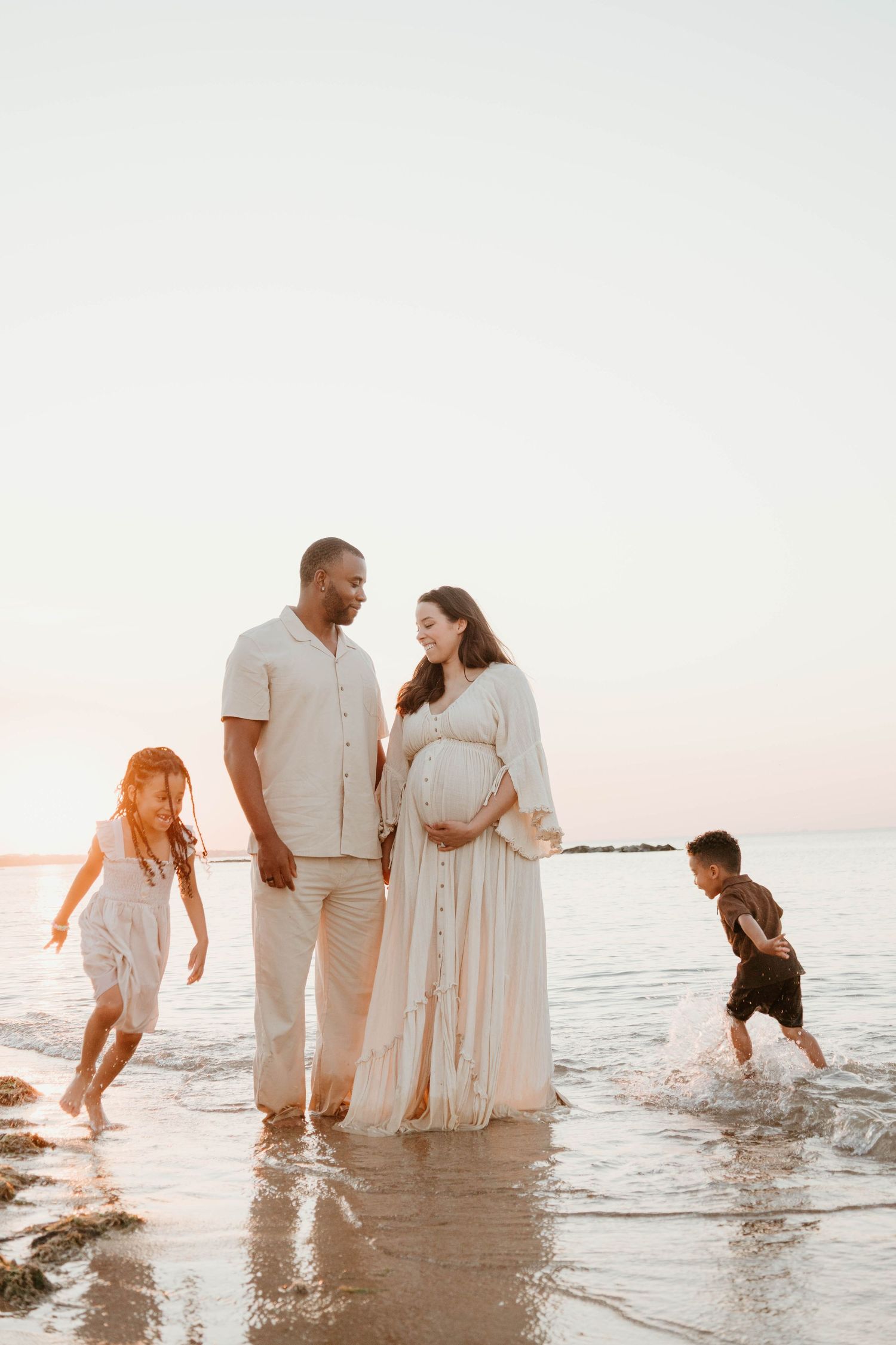 Pregnant woman in white dress with family on beach at sunset, children playing in waves.