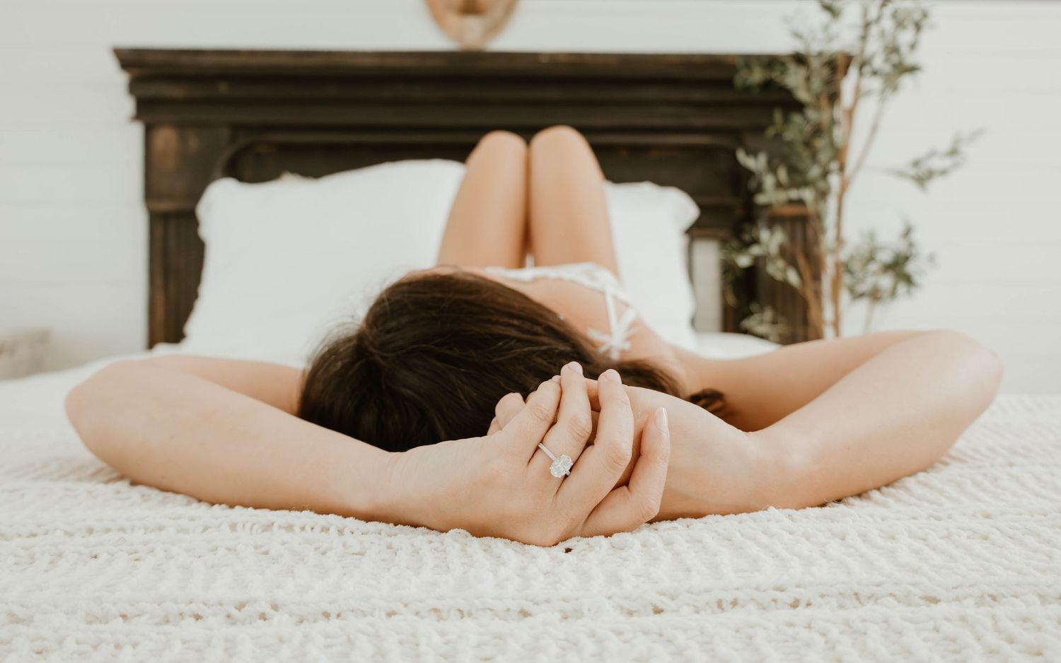 Woman lying face down on a white bed with hands clasped behind her head, wearing a ring, in a cozy bedroom.
