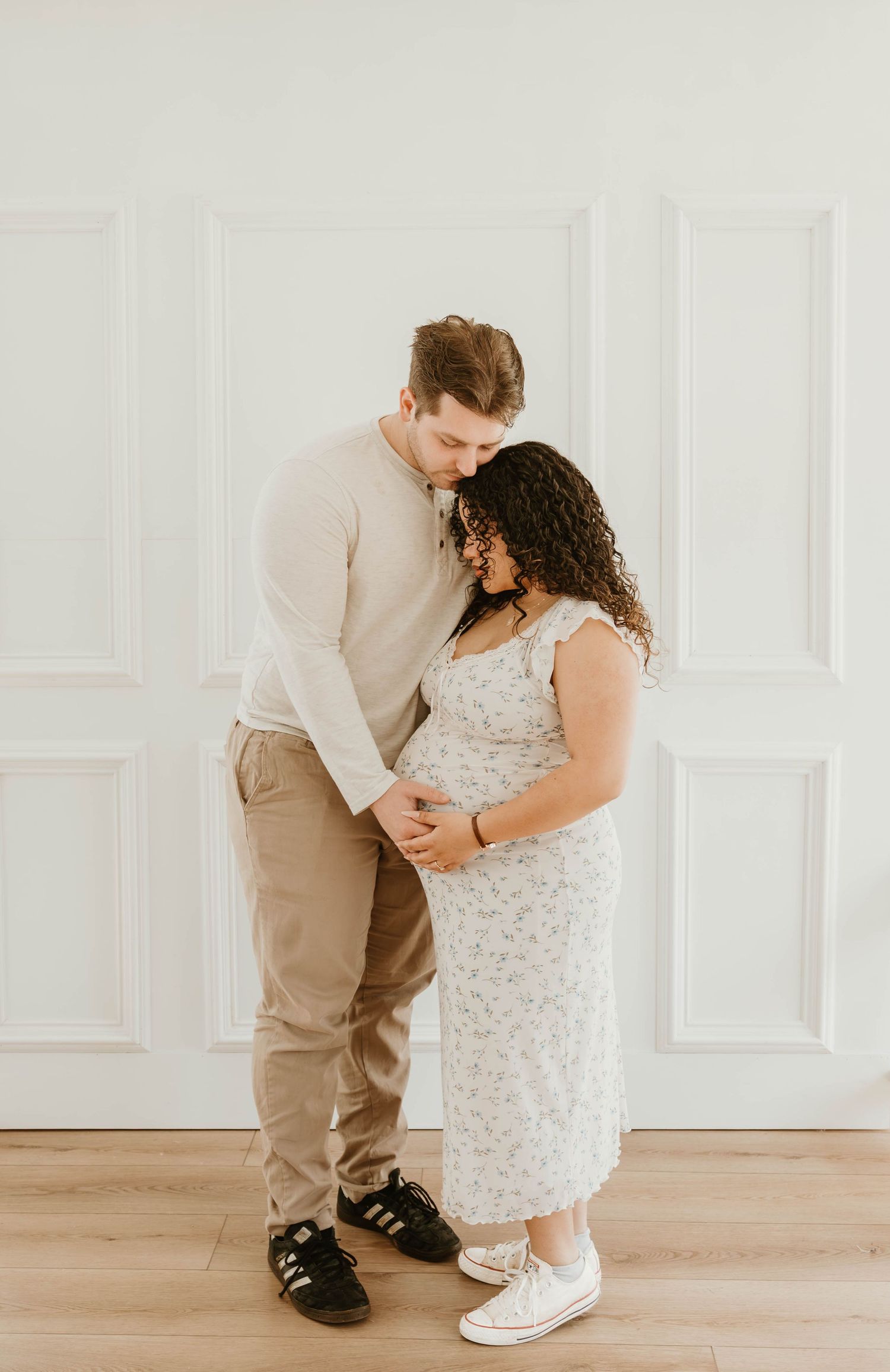 Pregnant couple in neutral tones sharing intimate moment, woman in white floral maternity dress holding baby bump.
