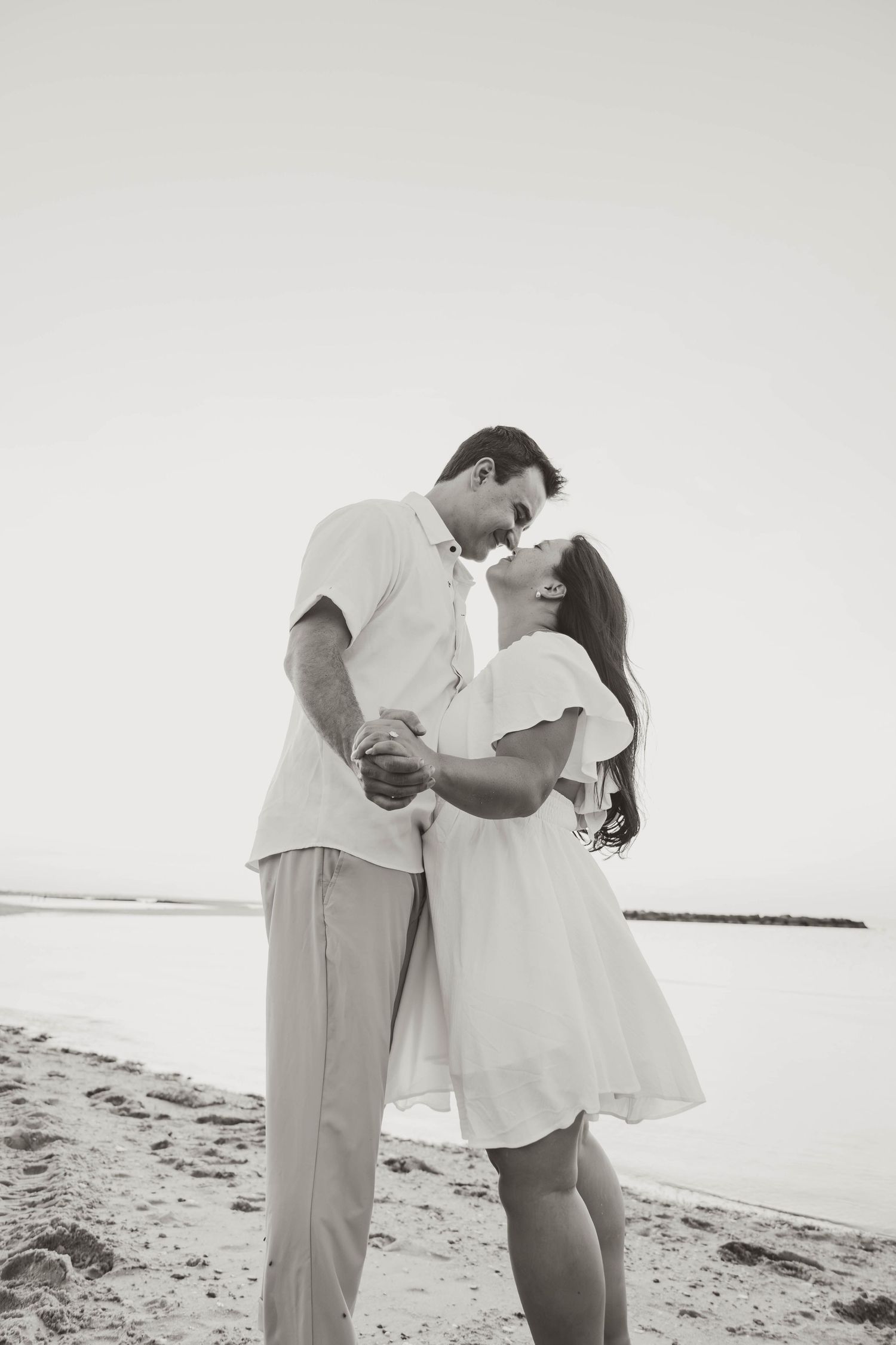 Black and white photo of a romantic couple kissing on a sandy beach, both dressed in white outfits.