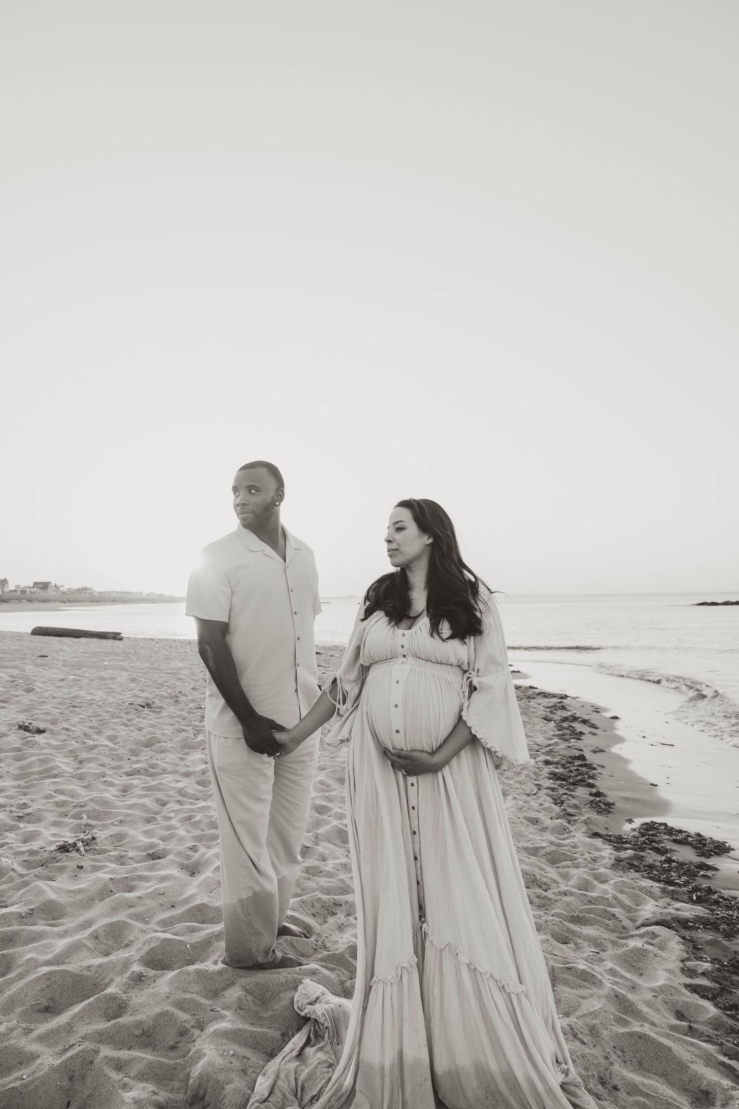 Black and white maternity photo of a couple on a sandy beach, pregnant woman in flowing dress holding her belly.
