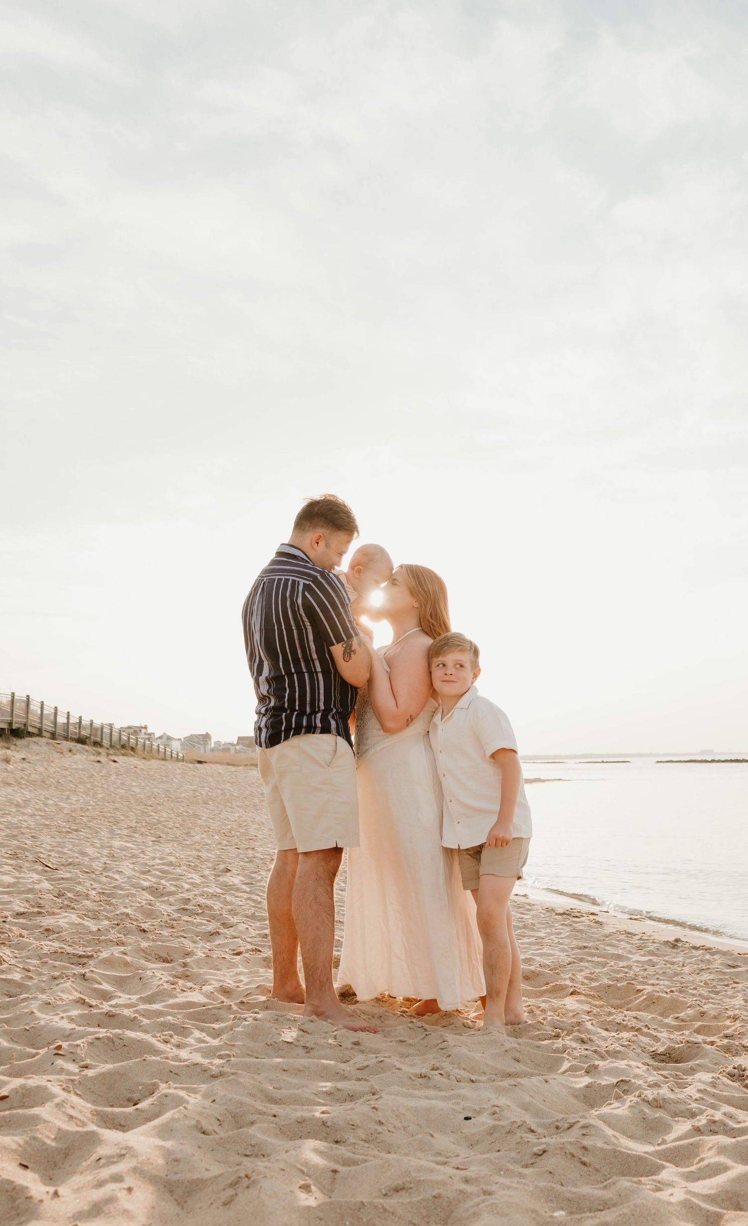 Family of three posing together on a sandy beach at golden hour, with ocean waves and a fence line in the background.