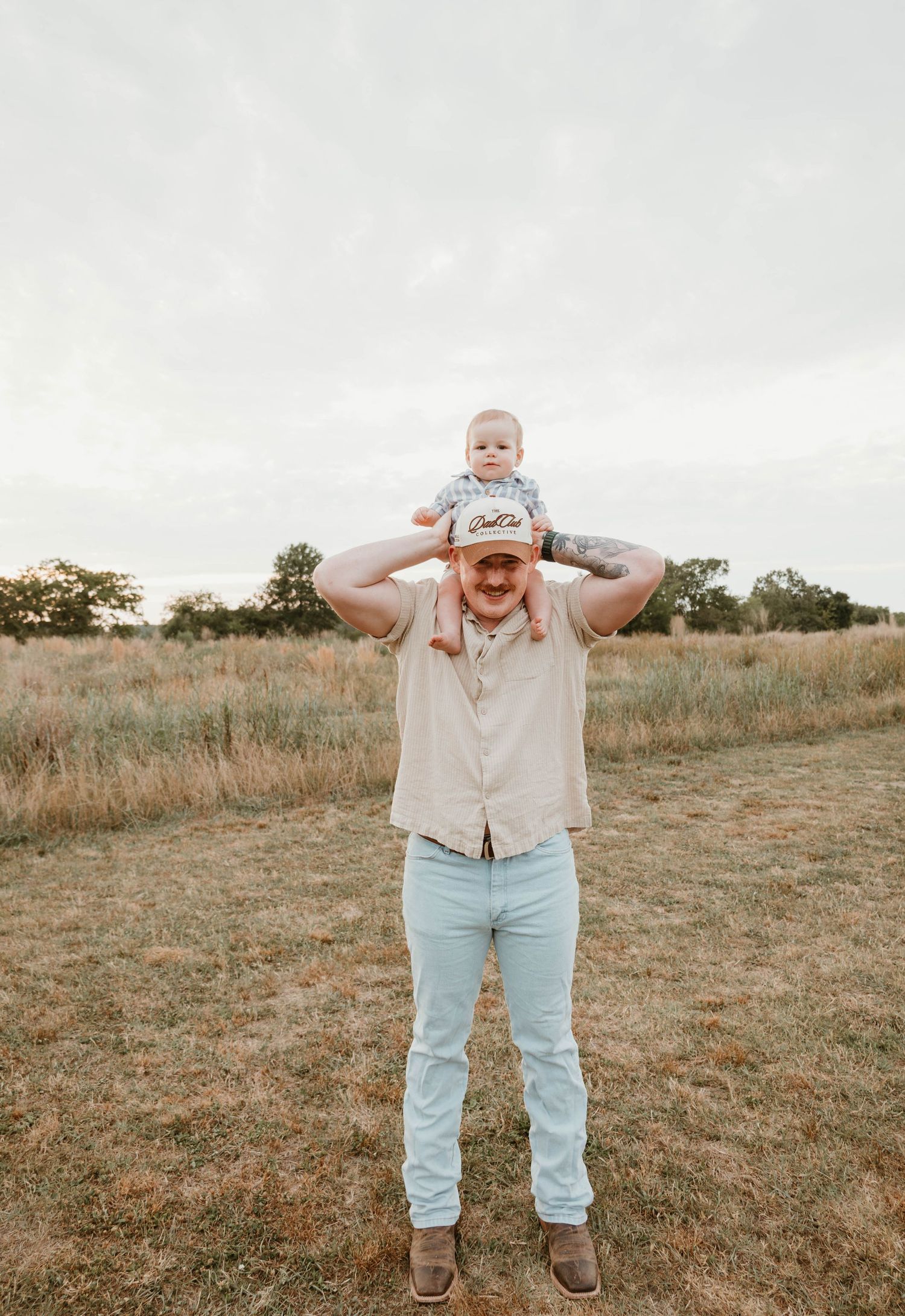 A tattooed man stands in a field holding a baby on his shoulders, smiling outdoors on a cloudy day.