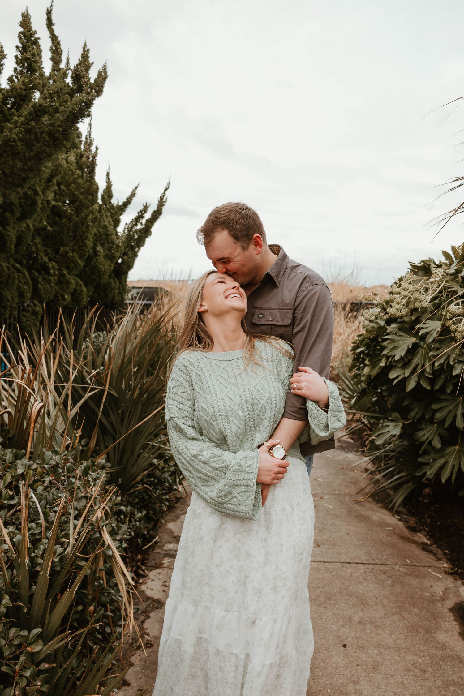 A romantic couple embraces on a garden path surrounded by tall greenery and shrubs during a cloudy day.