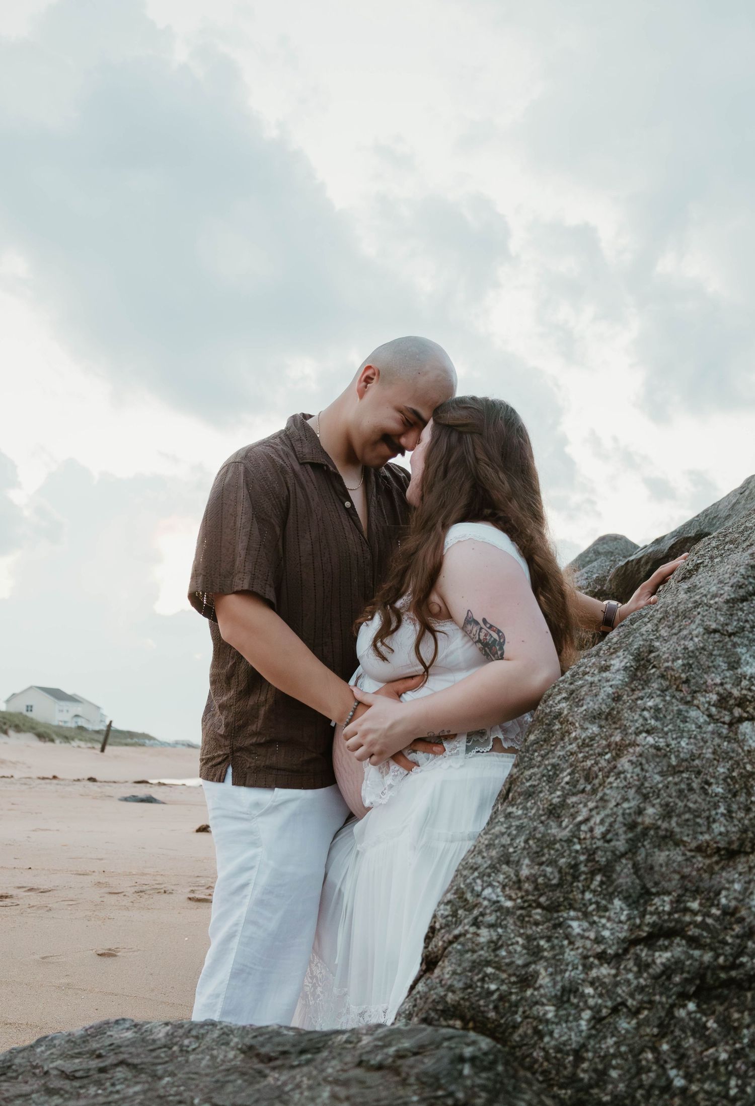 Couple sharing a romantic moment at the beach, woman in white dress showing baby bump, leaning against large rocks.