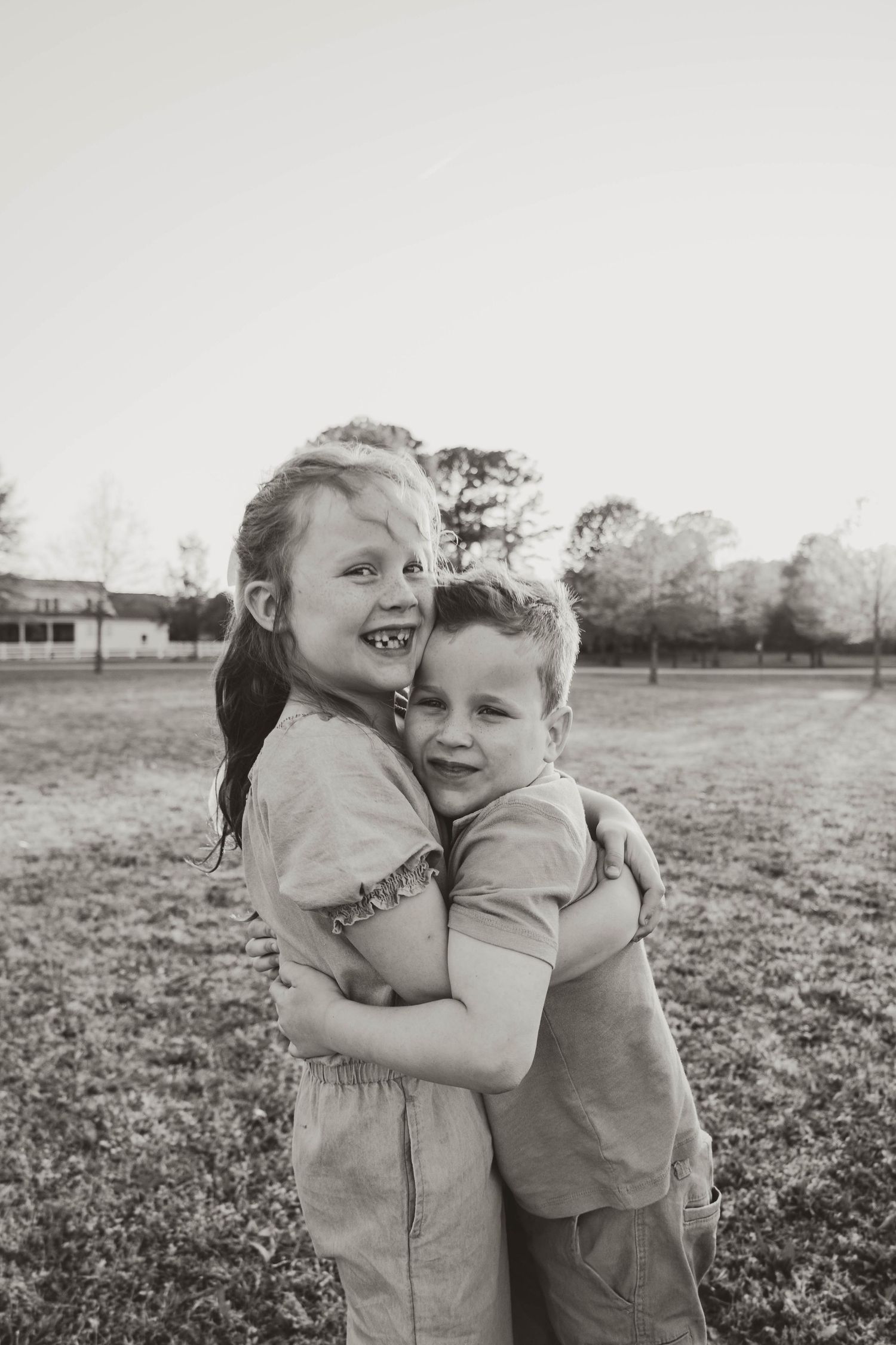Two young siblings share a warm hug in a black and white outdoor portrait.