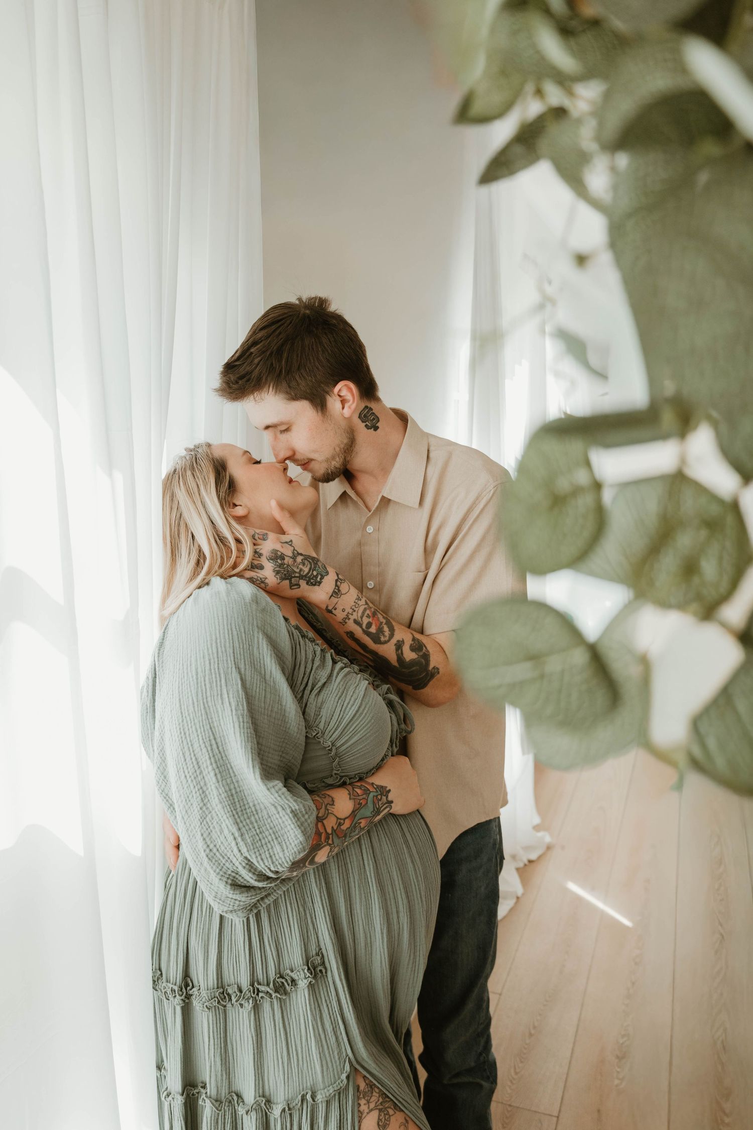 A couple embracing affectionately in a cozy indoor setting with greenery in the background.